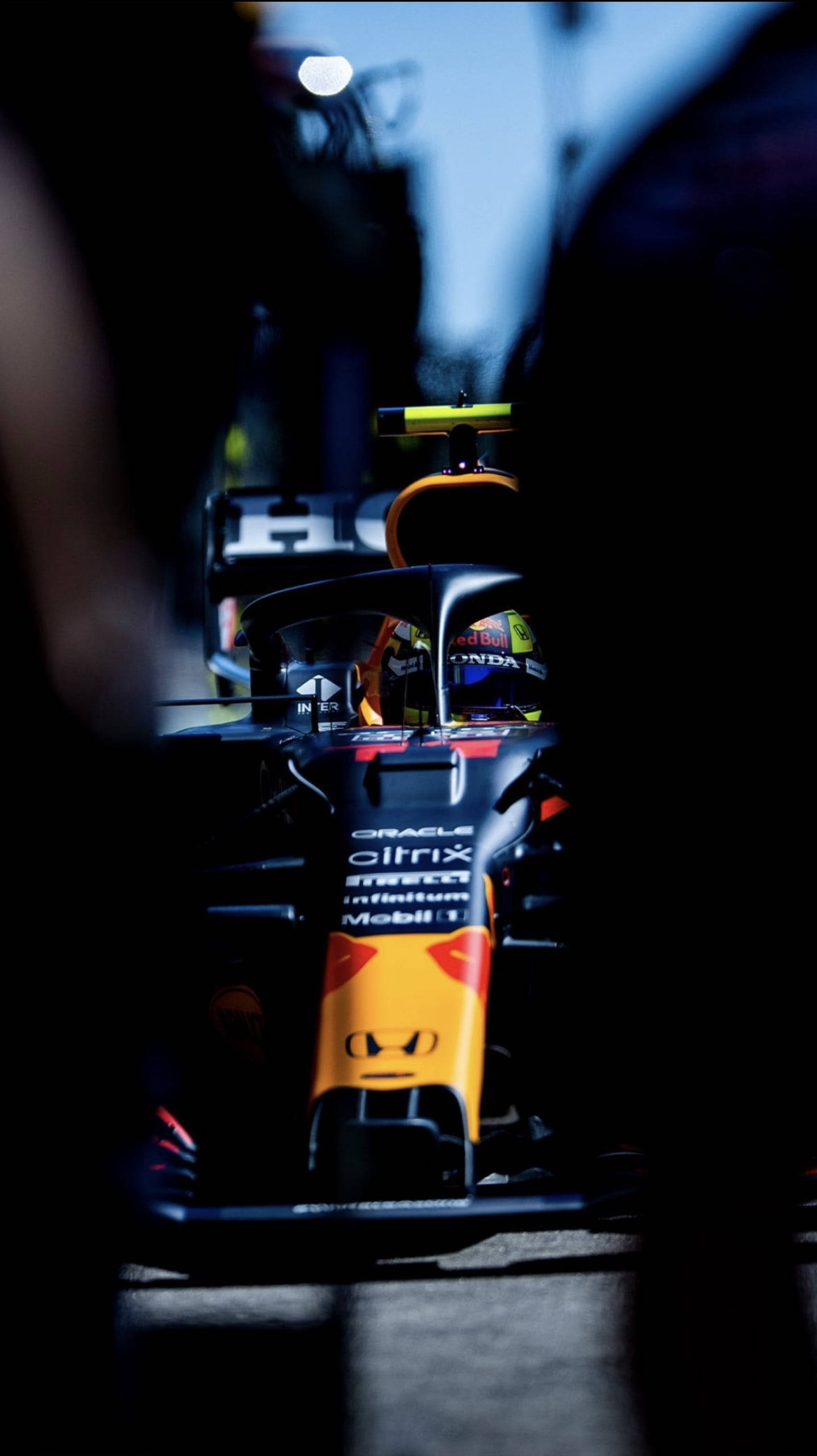 sergio perez 桌布 壁紙 — 一輛紅牛一級方程式賽車在昏暗的車庫中，在明亮的燈光下閃耀，前景有模糊的人影。