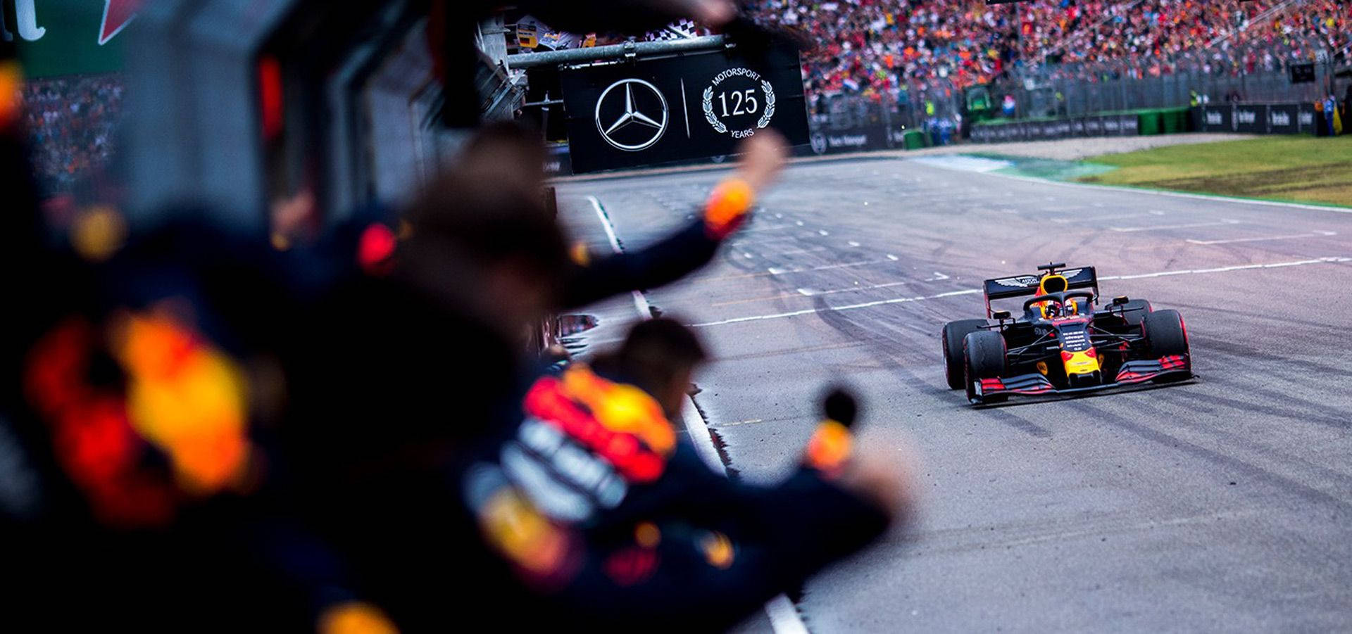 Verstappen 桌布 壁紙 — 一輛紅牛一級方程式賽車高速駛過在平治125周年紀念橫額前揮手的維修站人員。