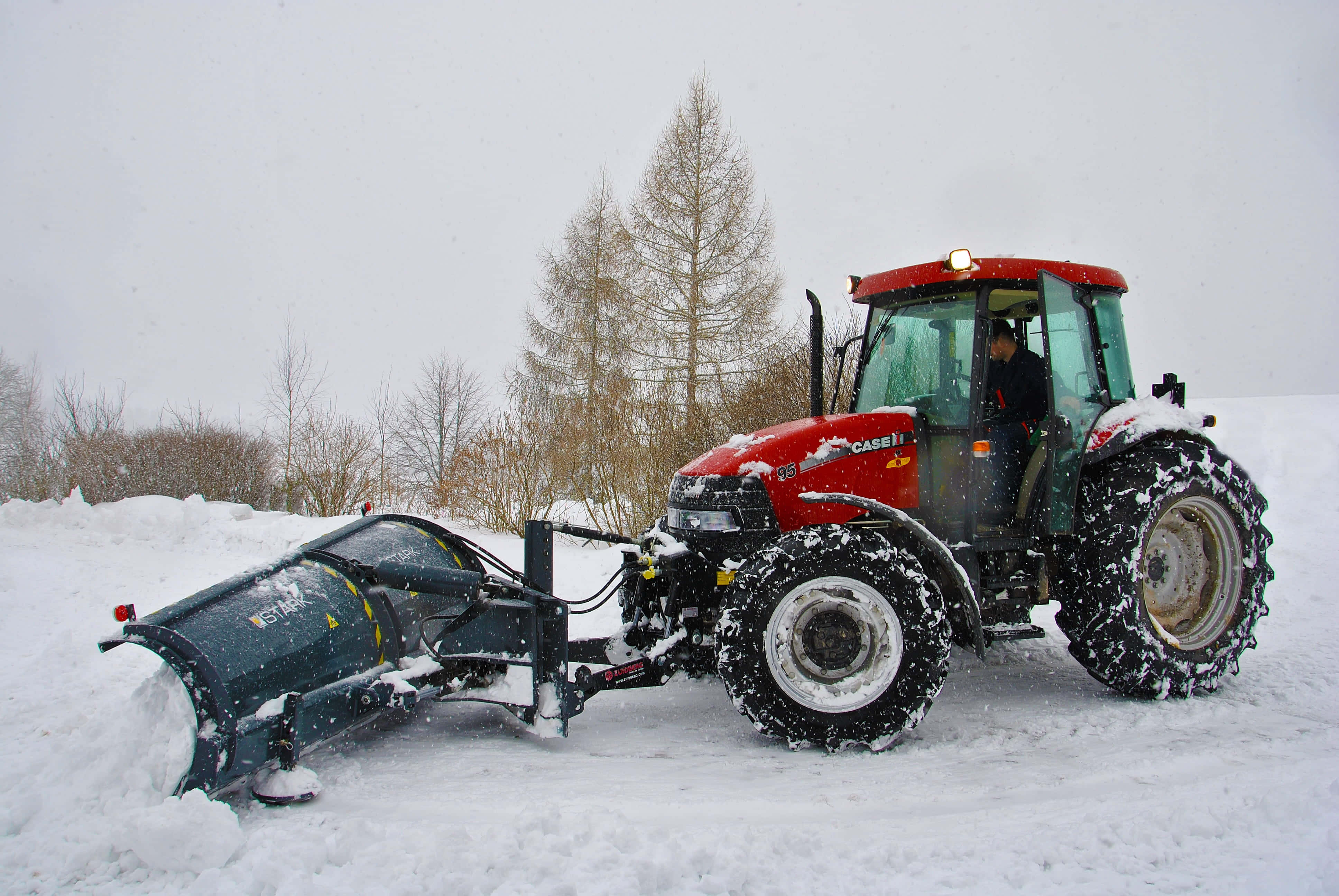 Snowplow 桌布 壁紙 — 一輛紅色Case拖拉機配備除雪機，在陰沉的冬日天空下清理積雪的鄉村道路，背景是光禿禿的樹木。
