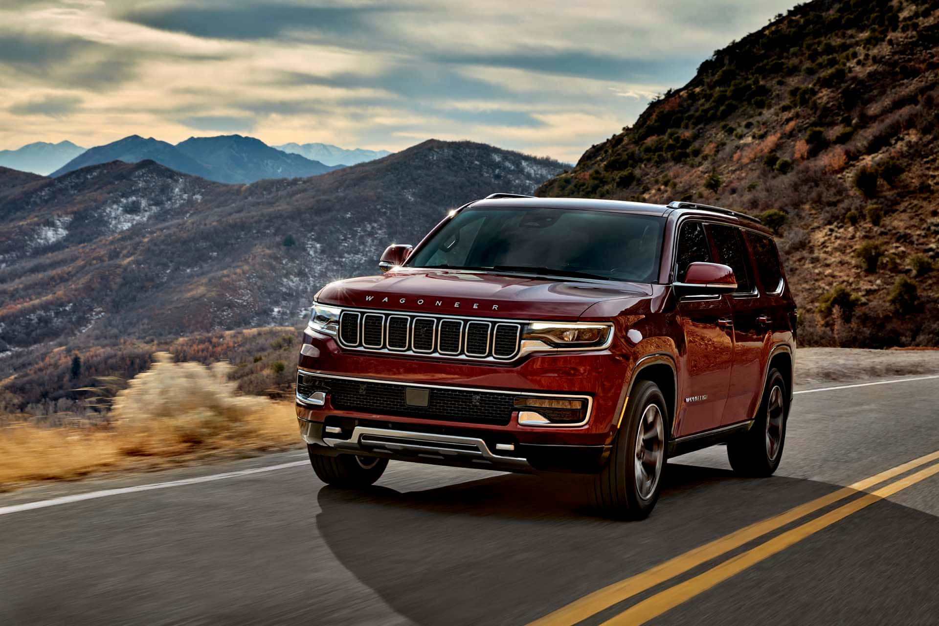 Jeep Wagoneer 桌布 壁紙 — 一輛紅色Jeep Wagoneer SUV在蜿蜒的山路上行駛，天空多雲，山頂覆蓋著積雪。