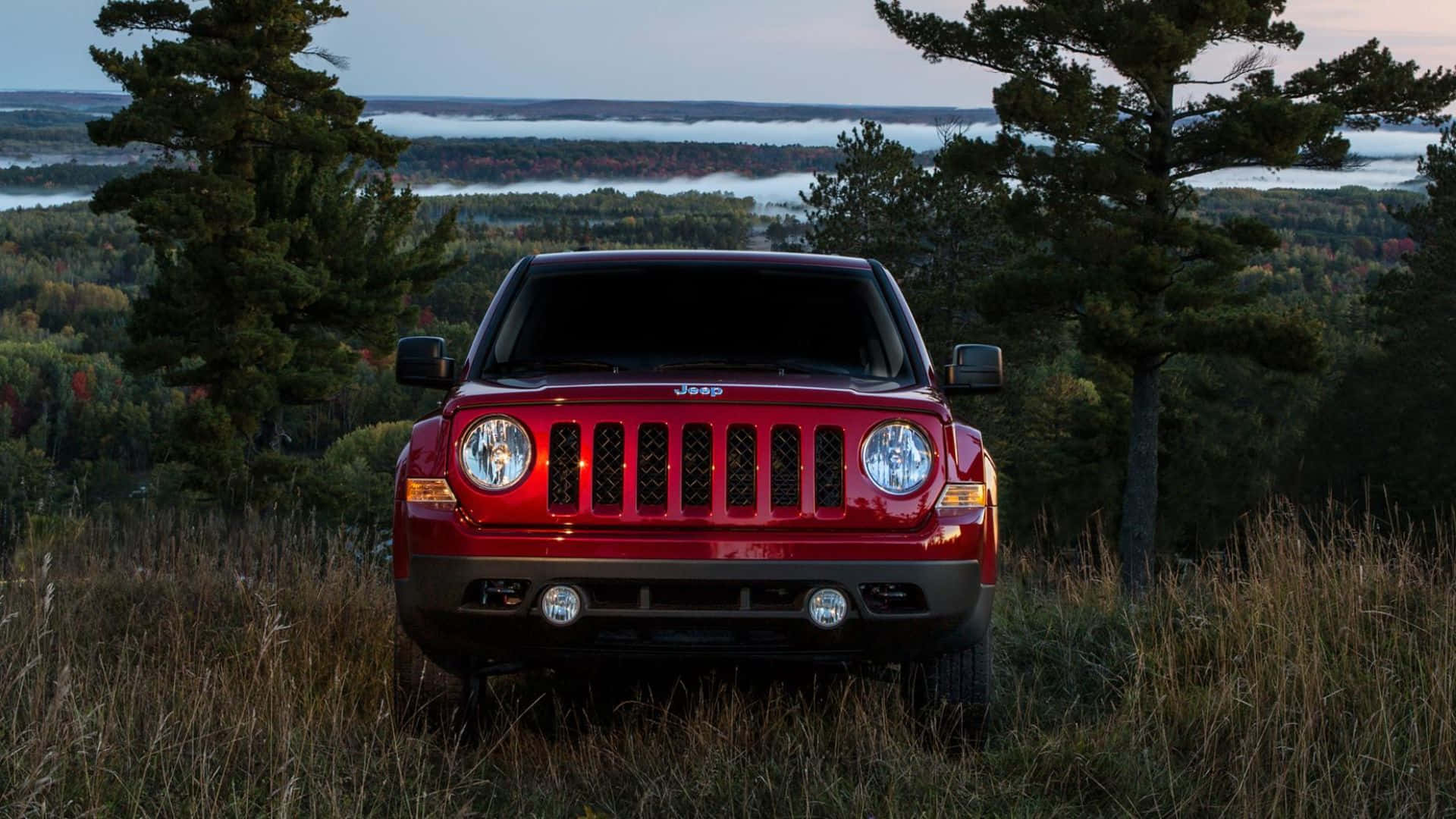 Jeep Patriot 桌布 壁紙 — 一輛紅色Jeep Wrangler在黎明時分面向前方，停在長滿草的山丘上，背後是霧氣瀰漫的森林和遠處的湖泊。