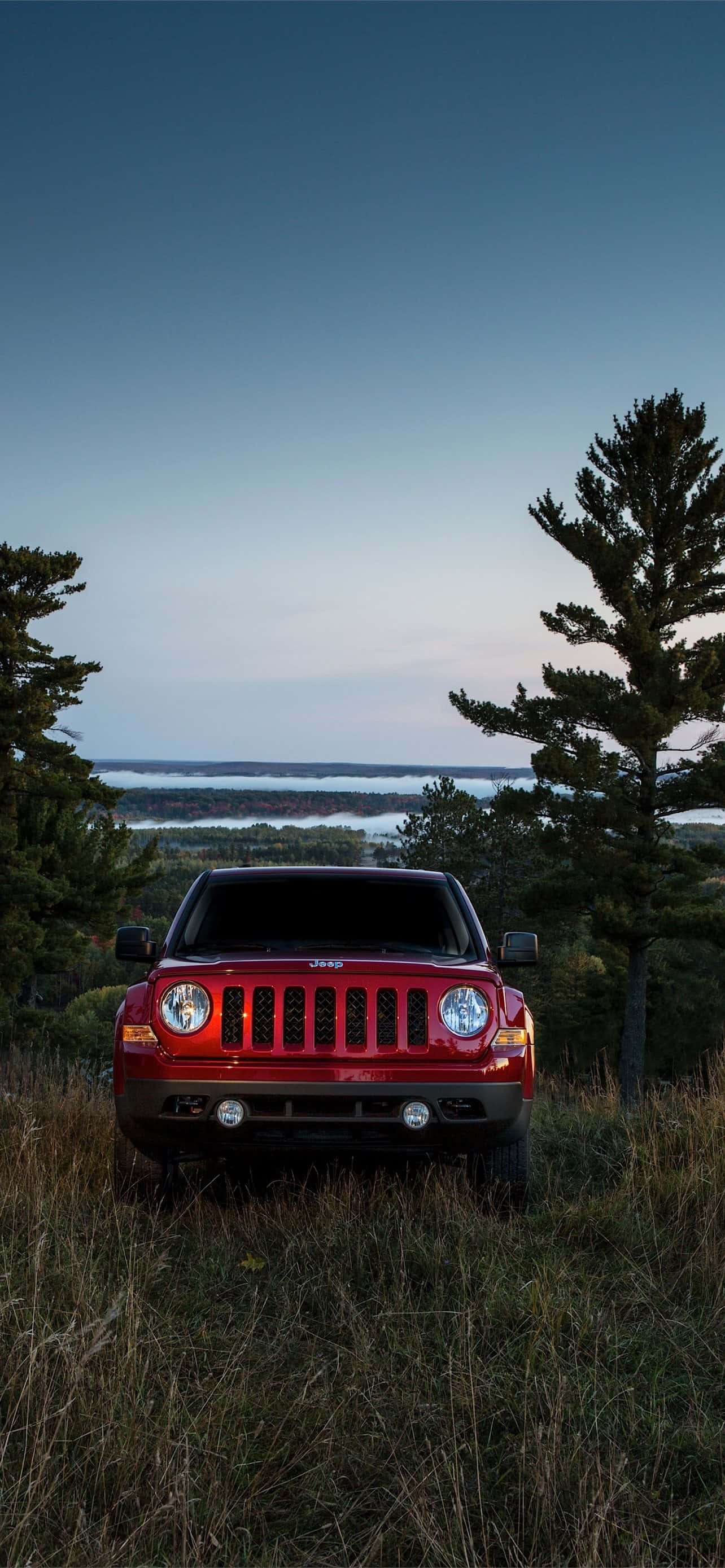 Jeep Patriot 桌布 壁紙 — 一輛紅色Jeep Wrangler在黃昏時分站在高高的草叢中，背景是湖泊和松樹。