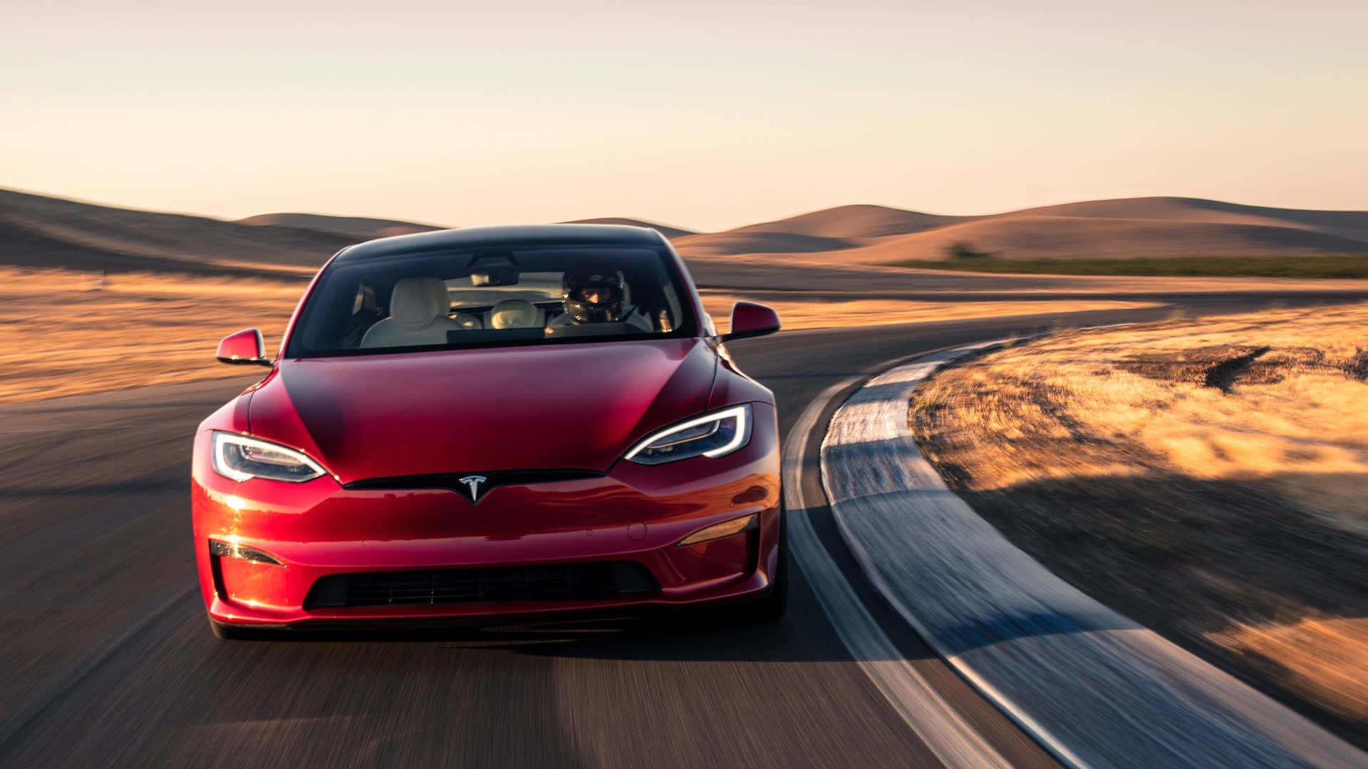 tesla 桌布 壁紙 — 一輛紅色Tesla Model S在日落時分沿著蜿蜒的沙漠公路飛馳，背景是起伏的沙丘。