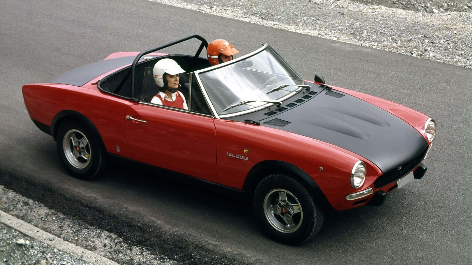 Abarth 桌布 壁紙 — 一輛紅色和黑色的 Fiat 124 Spider 開篷車，載著兩位戴頭盔的司機，在鋪設的道路上行駛。