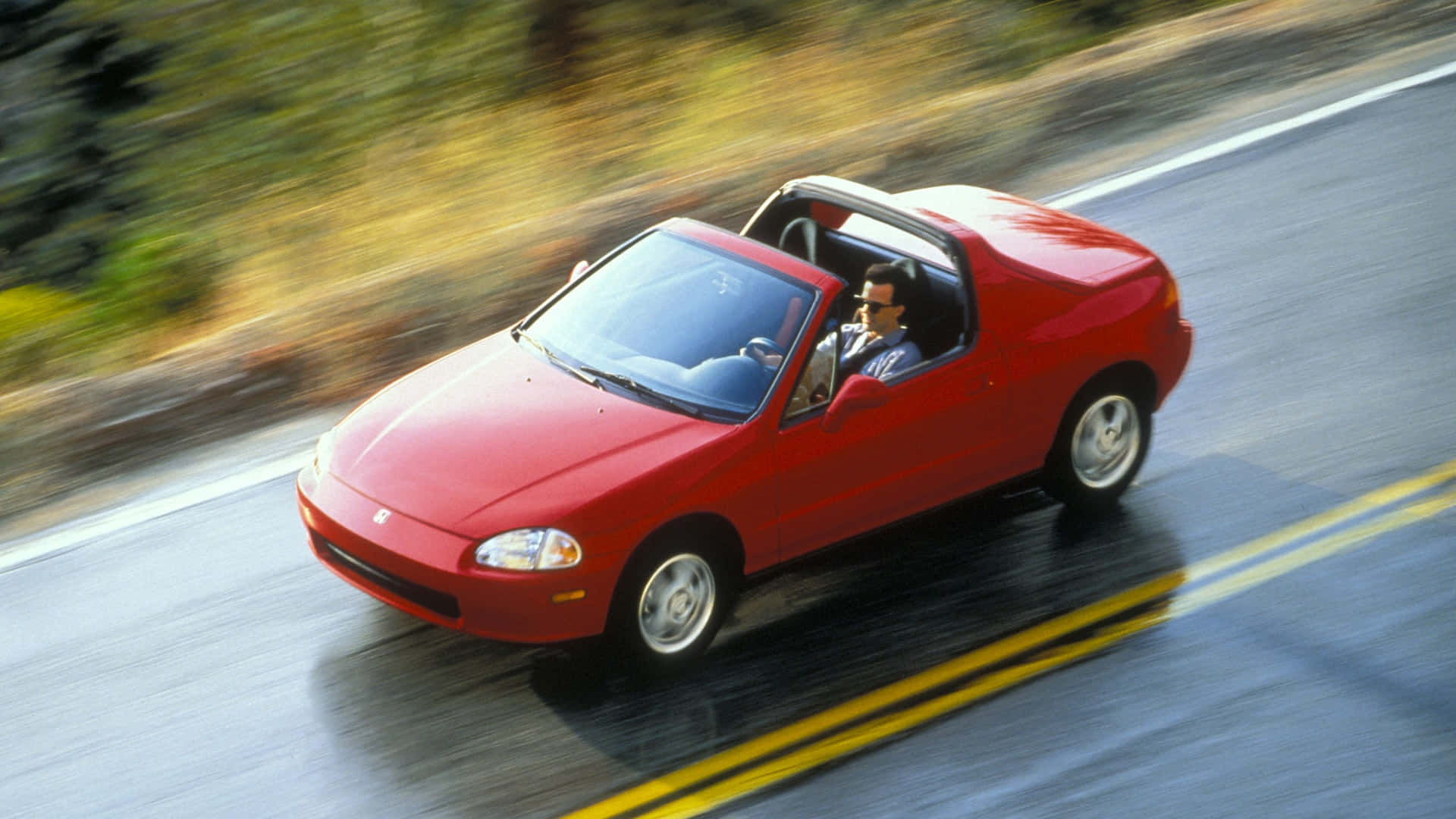 Honda Civic Del Sol 桌布 壁紙 — 一輛紅色本田Miata開篷跑車在蜿蜒的道路上疾馳，周圍是模糊的綠化和黃線。