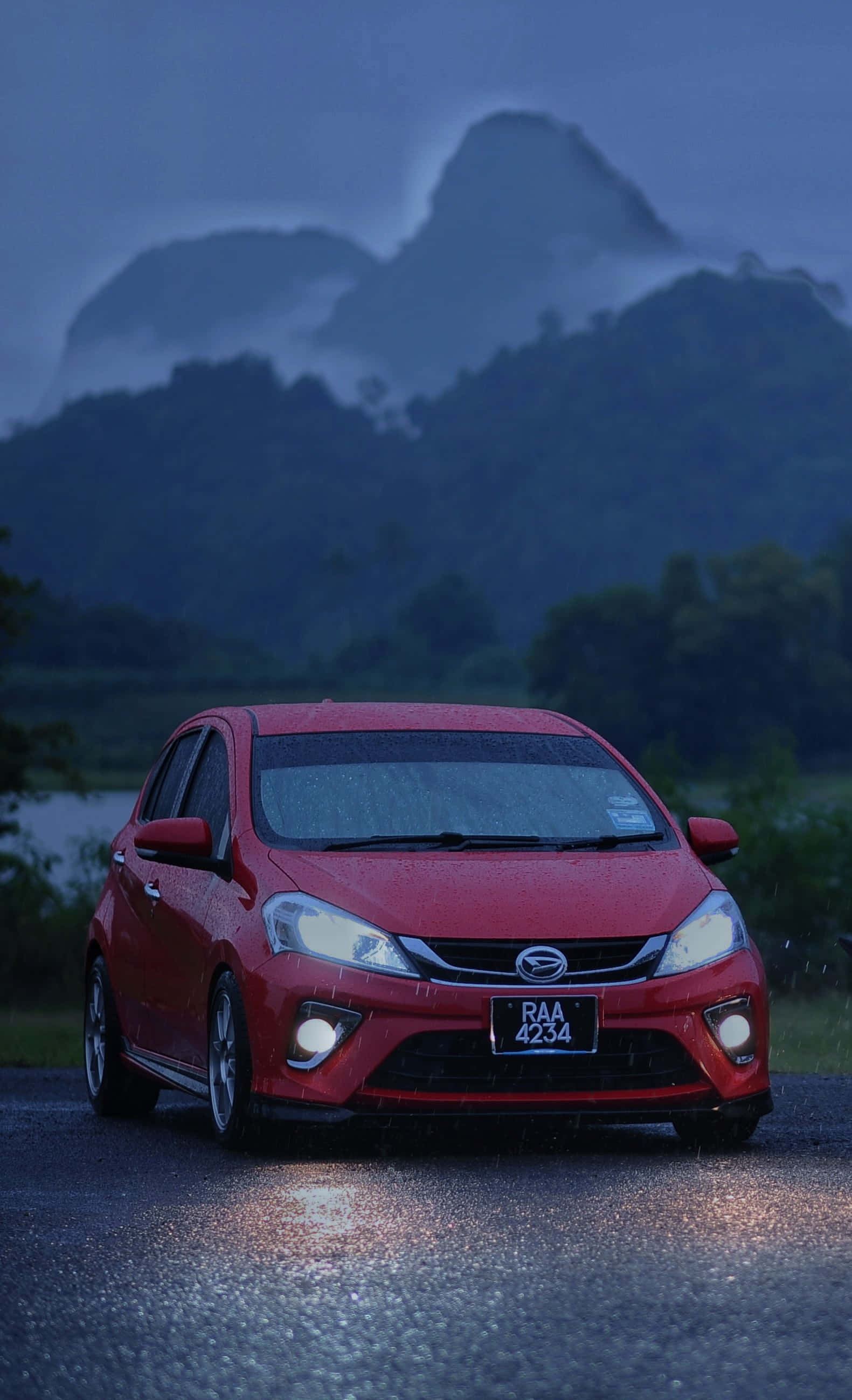 Daihatsu 桌布 壁紙 — 一輛紅色汽車亮著車頭燈，在黃昏時分霧氣瀰漫的山巒天空下行駛在濕滑的道路上。
