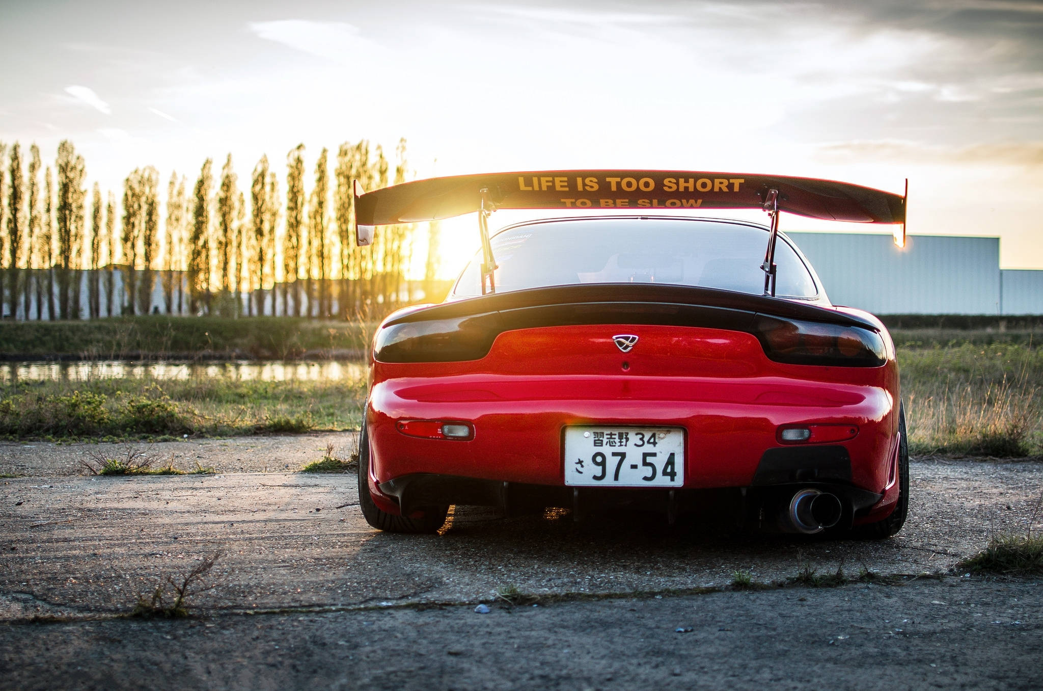 Mazda 桌布 壁紙 — 一輛紅色萬事得RX-7，帶有大型尾翼和標語「LIFE IS TOO SHORT TO BE SLOW」，停泊在日落時分龜裂的瀝青路上。