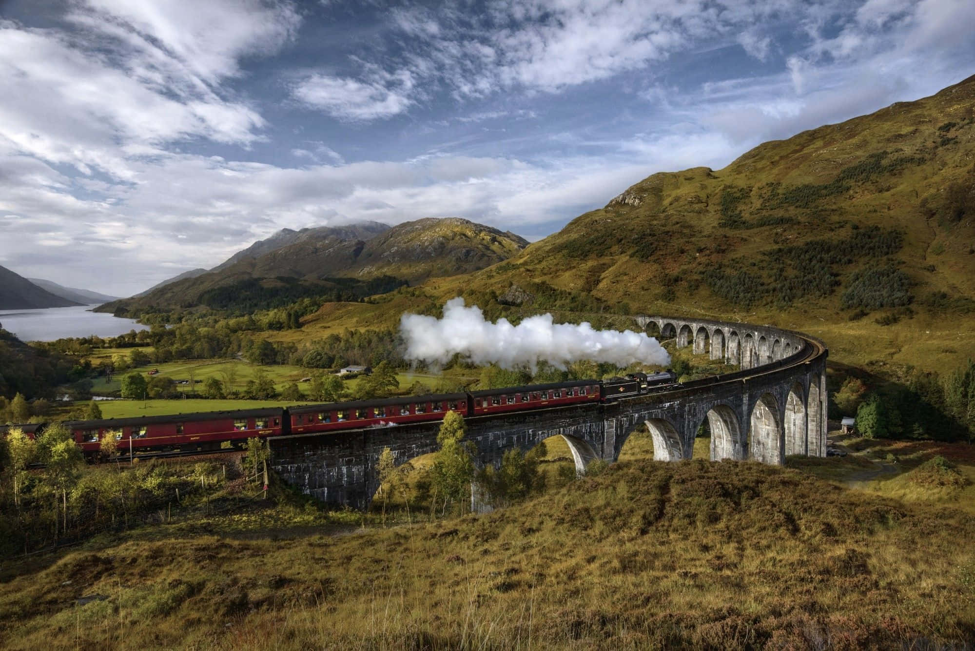 The Hogwarts Express Train 桌布 壁紙 — 一輛紅色蒸汽火車在部分多雲的天空下，穿過崎嶇的綠色山谷，沿著石拱橋彎曲行駛，噴出濃厚的白色煙霧，山谷中有山脈和湖泊。
