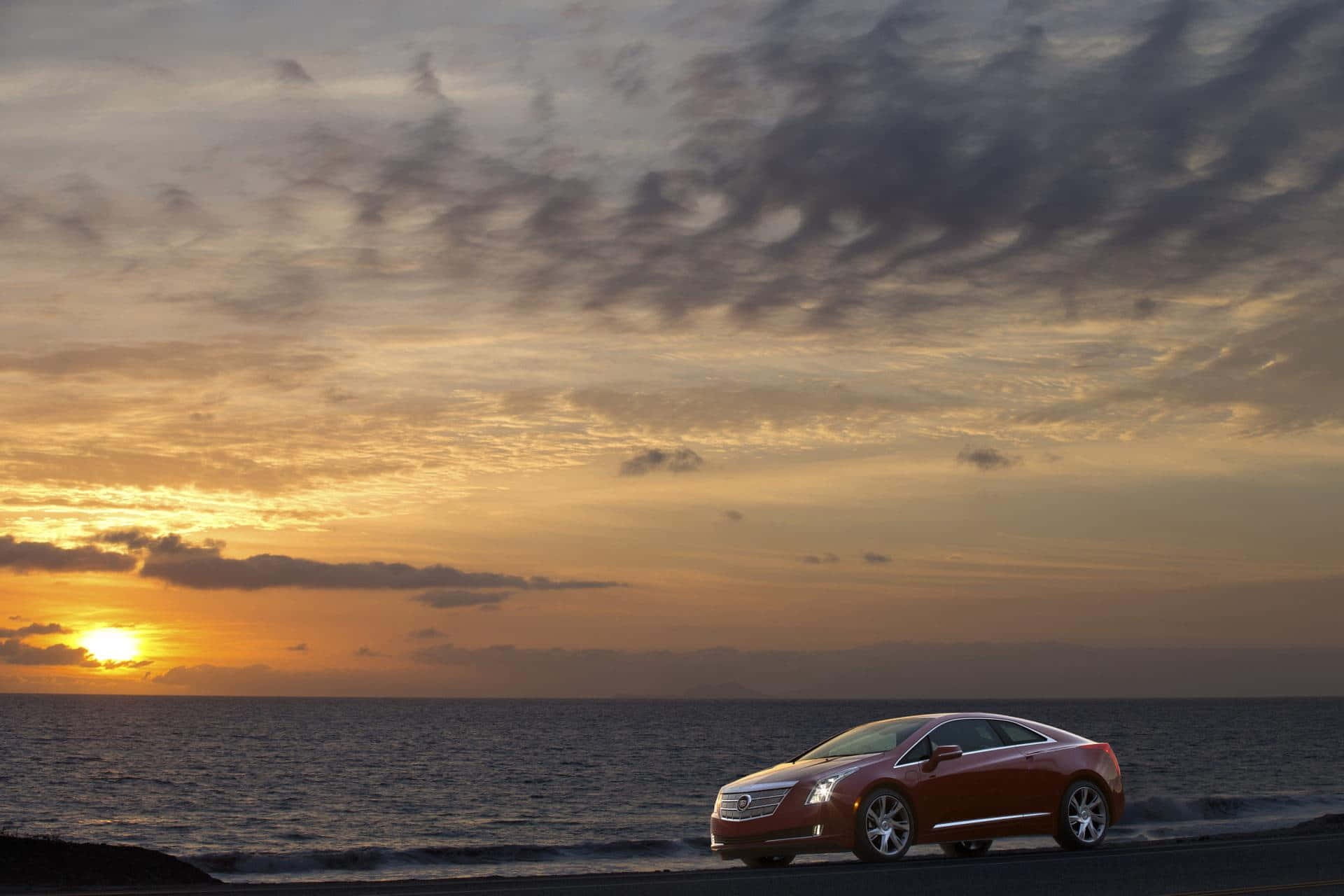 Cadillac Elr 桌布 壁紙 — 一輛紅色豪華轎跑車停泊在日落時分的海岸公路上，戲劇性的雲彩和金色光芒映照在海面上。
