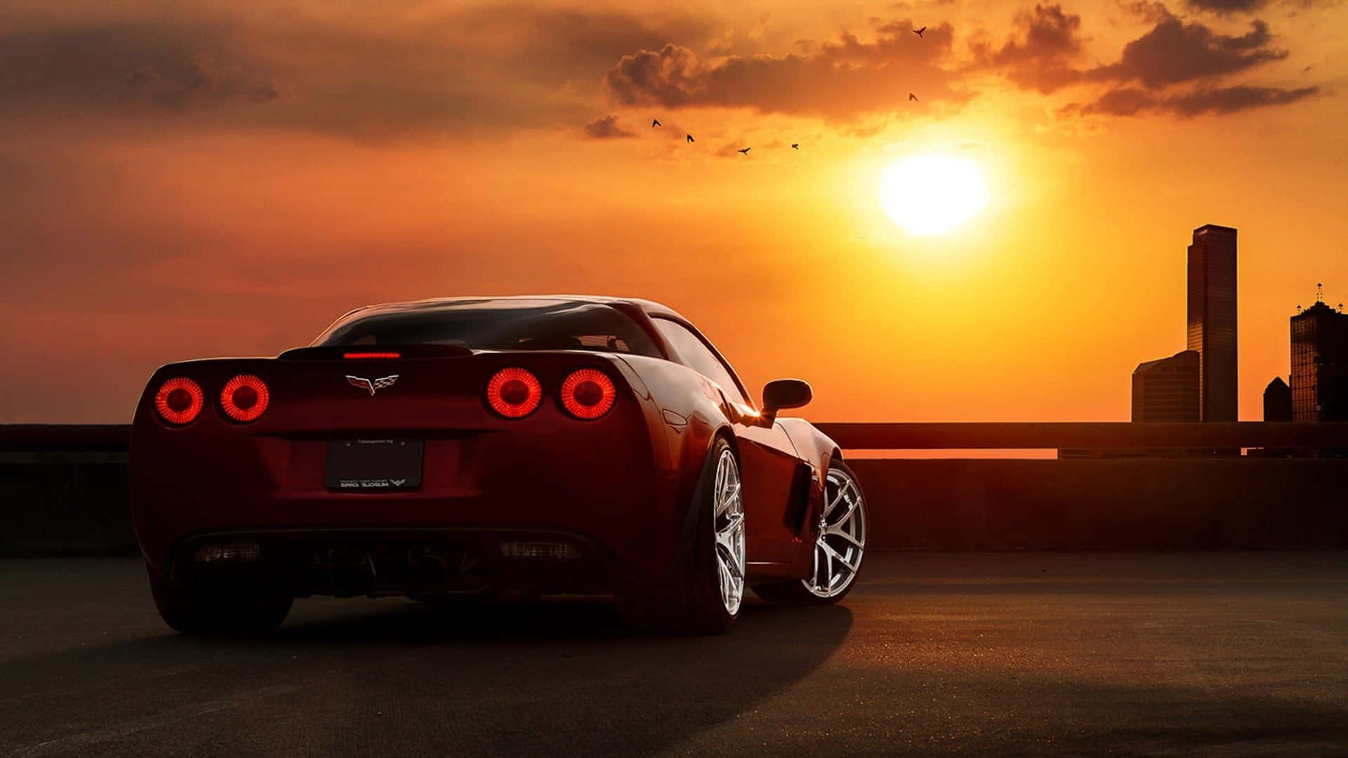 Chevrolet Corvette Z06 桌布 壁紙 — 一輛紅色跑車亮著尾燈停在高速公路天橋上，夕陽在城市的輪廓後落下，鳥兒在頭頂飛翔。