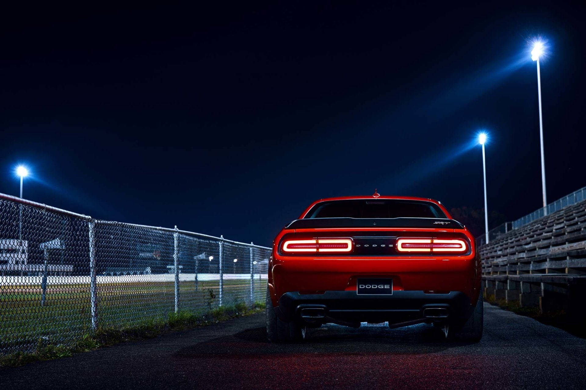 Mopar 2022 Dodge Challenger 壁紙 — 一輛紅色道奇挑戰者在夜間停放在明亮的體育場燈光下，背景有鐵絲網圍欄和看台。