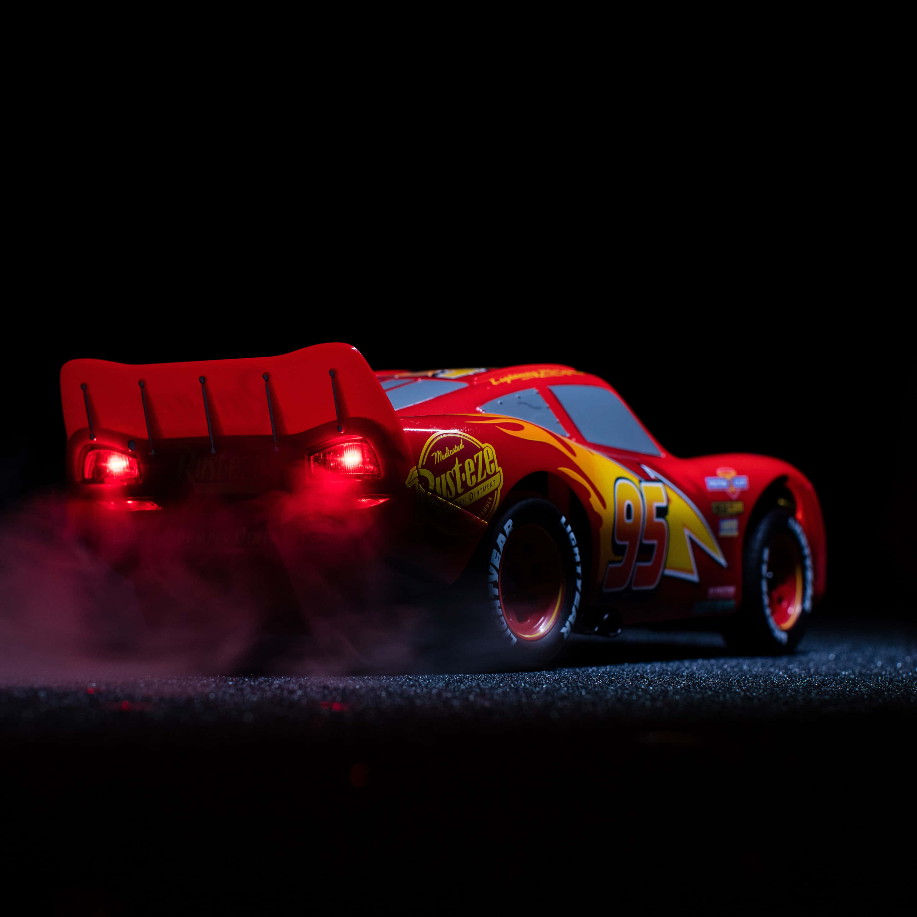 Disney Cars 桌布 壁紙 — 一輛紅色閃電王麥坤玩具車，帶有發光尾燈和煙霧，背景為黑色。