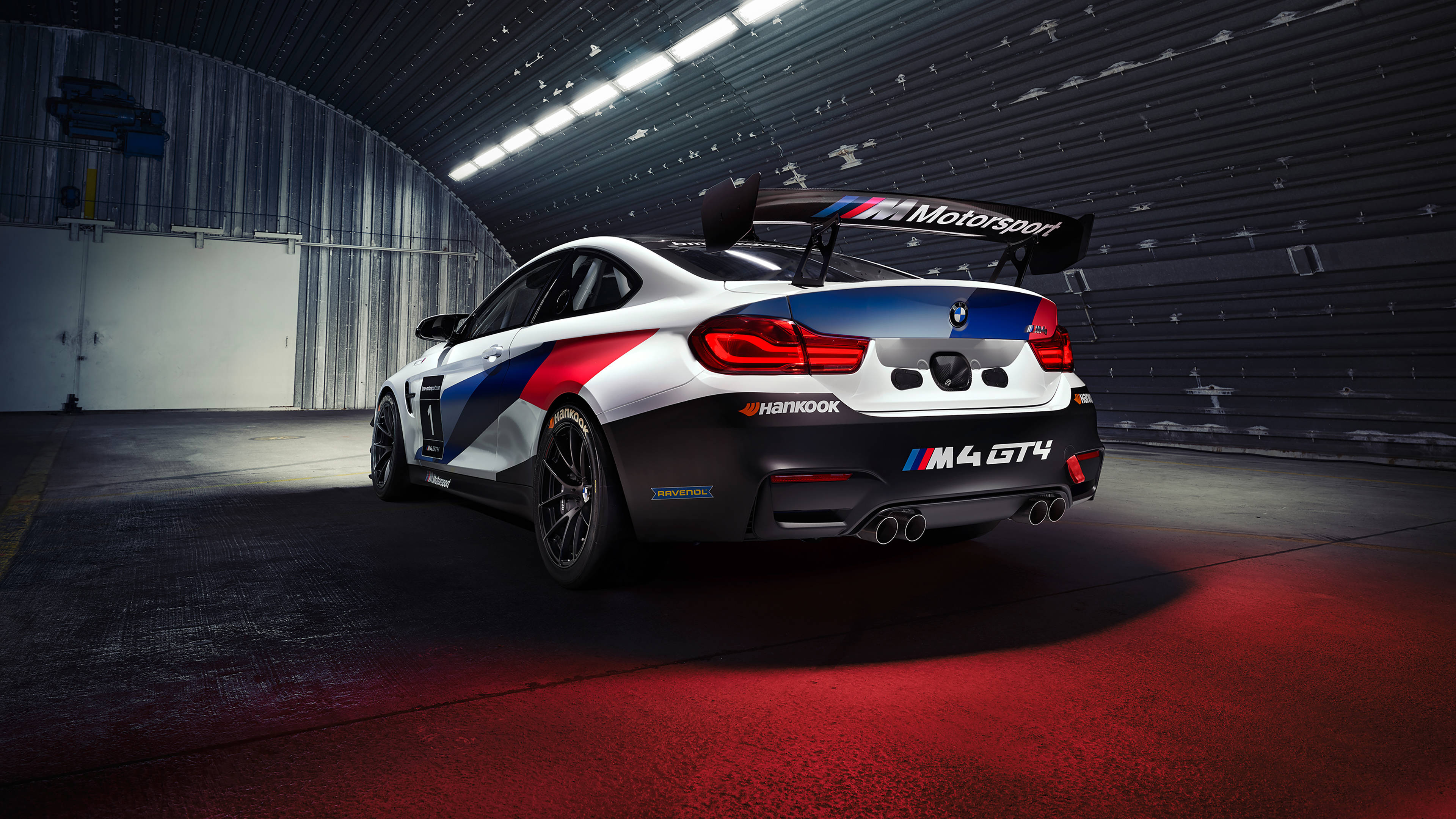 Bmw 桌布 壁紙 — 一輛紅藍白塗裝的BMW M4 GT4賽車停放在光線昏暗、帶有紅色地板照明的車庫中。