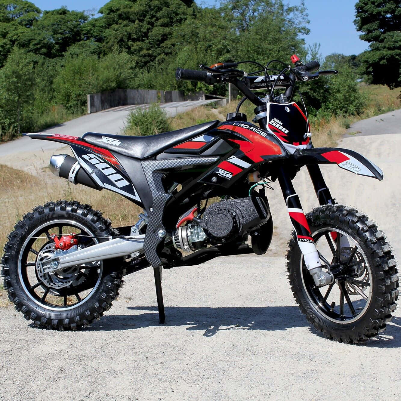 Dirtbike 桌布 壁紙 — 一輛紅黑白色的越野電單車，配備防滑輪胎，停在碎石路上，周圍綠樹成蔭，天空清澈湛藍。