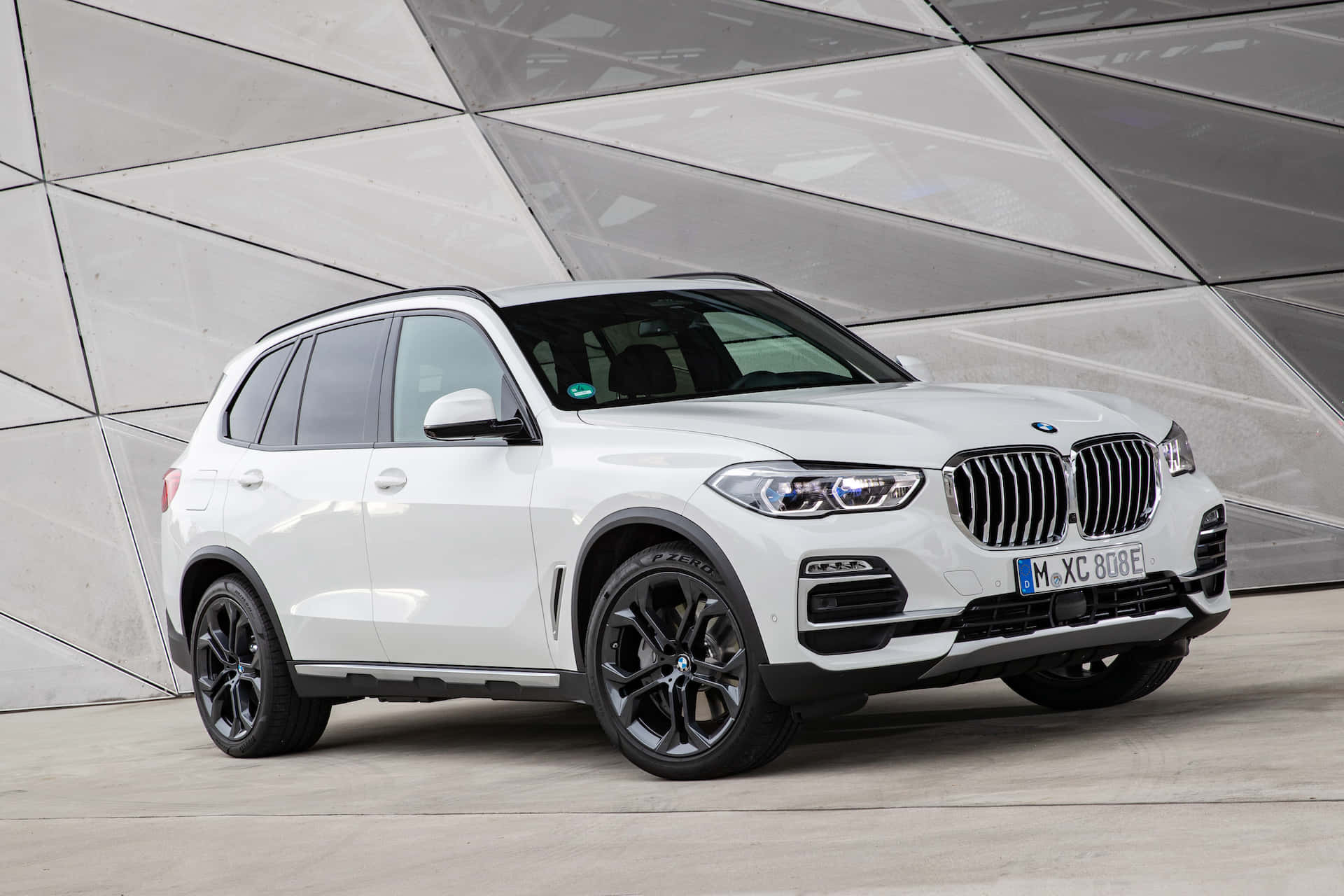 Bmw X5 桌布 壁紙 — 一輛純白色BMW X5 SUV配黑色輪圈，自信地矗立在現代幾何玻璃與混凝土建築外牆前。