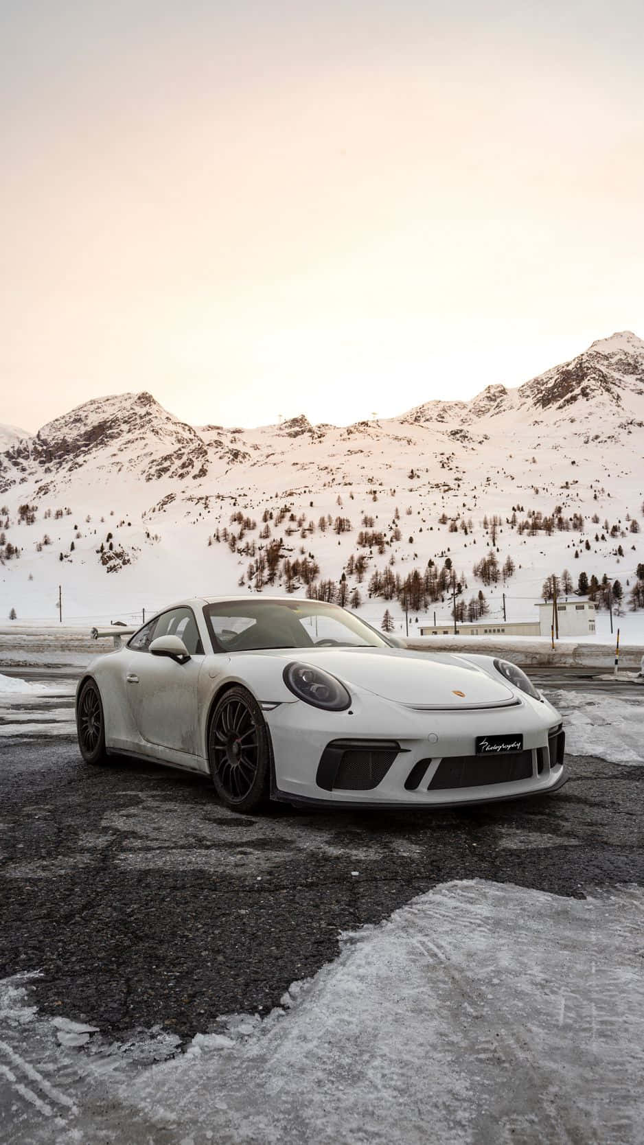 Porsche iPhone 桌布 壁紙 — 一輛純白色的保時捷911 GT3停在結冰的瀝青路上，背景是白雪覆蓋的山脈和光禿禿的樹木，天空呈現柔和的粉紅色。
