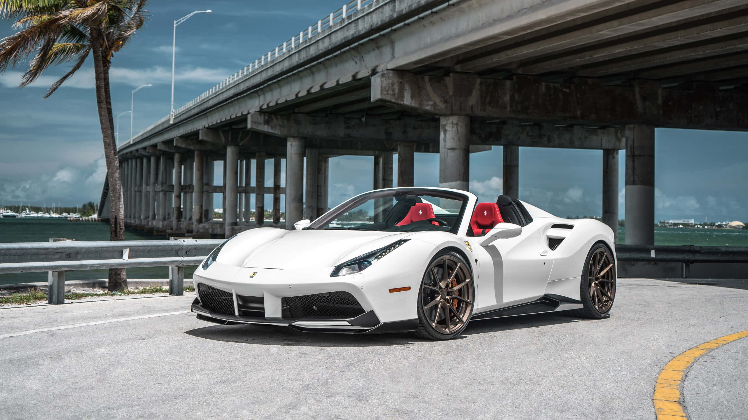 Ferrari 488 Spider 桌布 壁紙 — 一輛純白色的法拉利跑車，配備紅色座椅，停泊在混凝土橋下，旁邊有棕櫚樹和水。