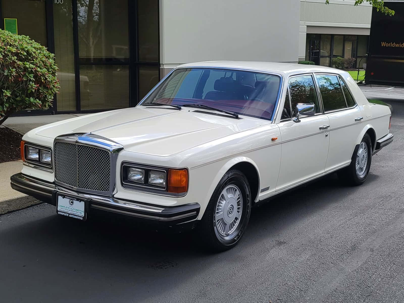 Bentley Eight 桌布 壁紙 — 一輛純白色的賓利轎車在日光下閃耀，停在瀝青路面上，旁邊是帶有玻璃窗和綠植的現代建築。