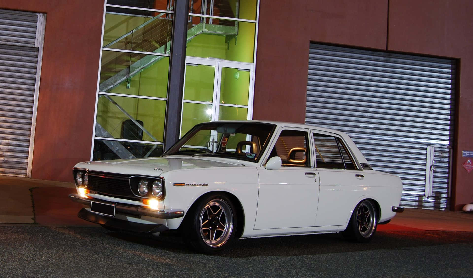 Datsun 桌布 壁紙 — 一輛純白色經典房車配備訂製輪圈，在夜燈下閃閃發光，旁邊是一座帶有綠色發光窗戶的現代建築。