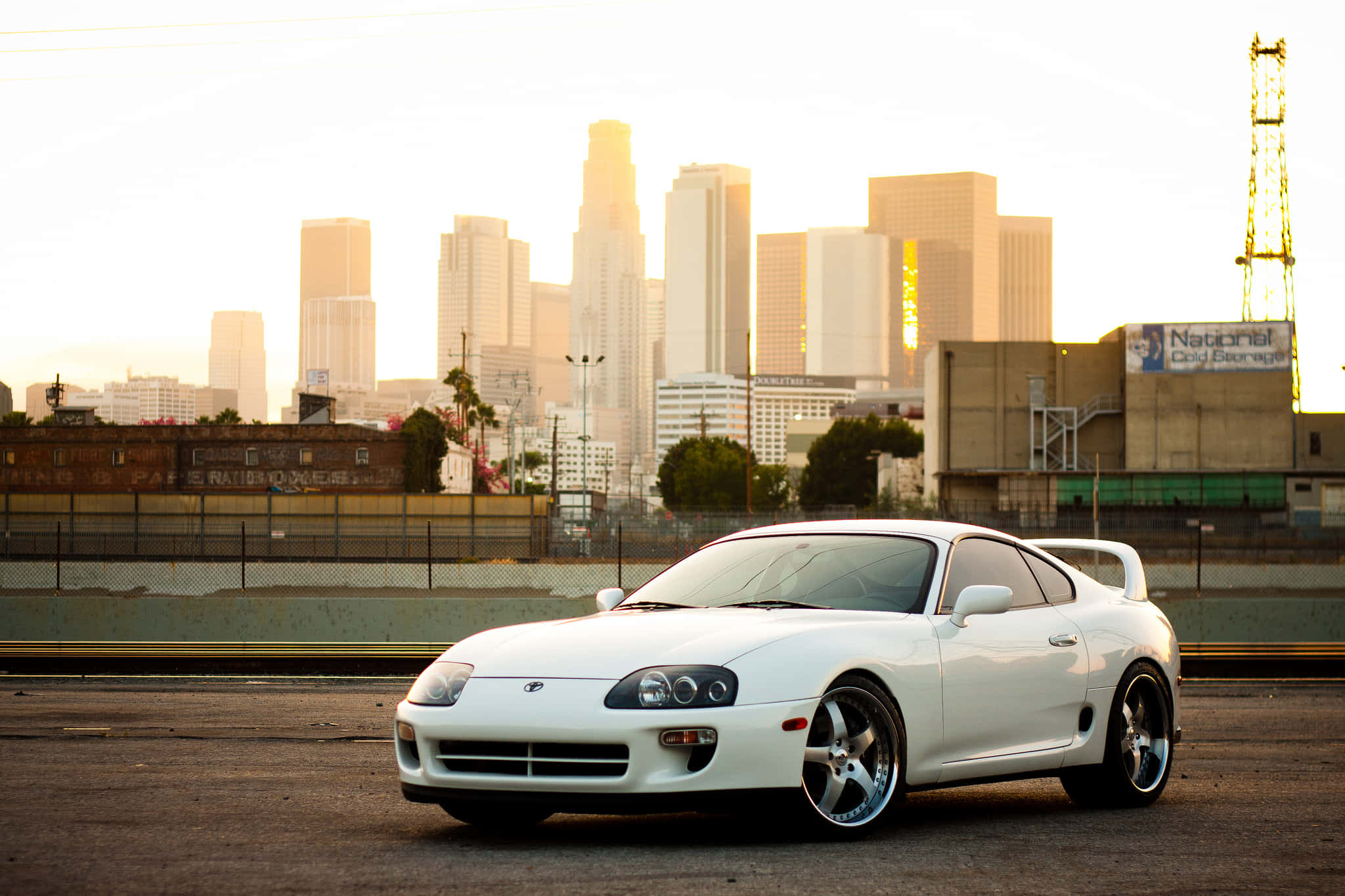 Supra 桌布 壁紙 — 一輛純白色跑車在金色夕陽光下閃閃發光，背景是朦朧的城市天際線和高聳的摩天大樓。