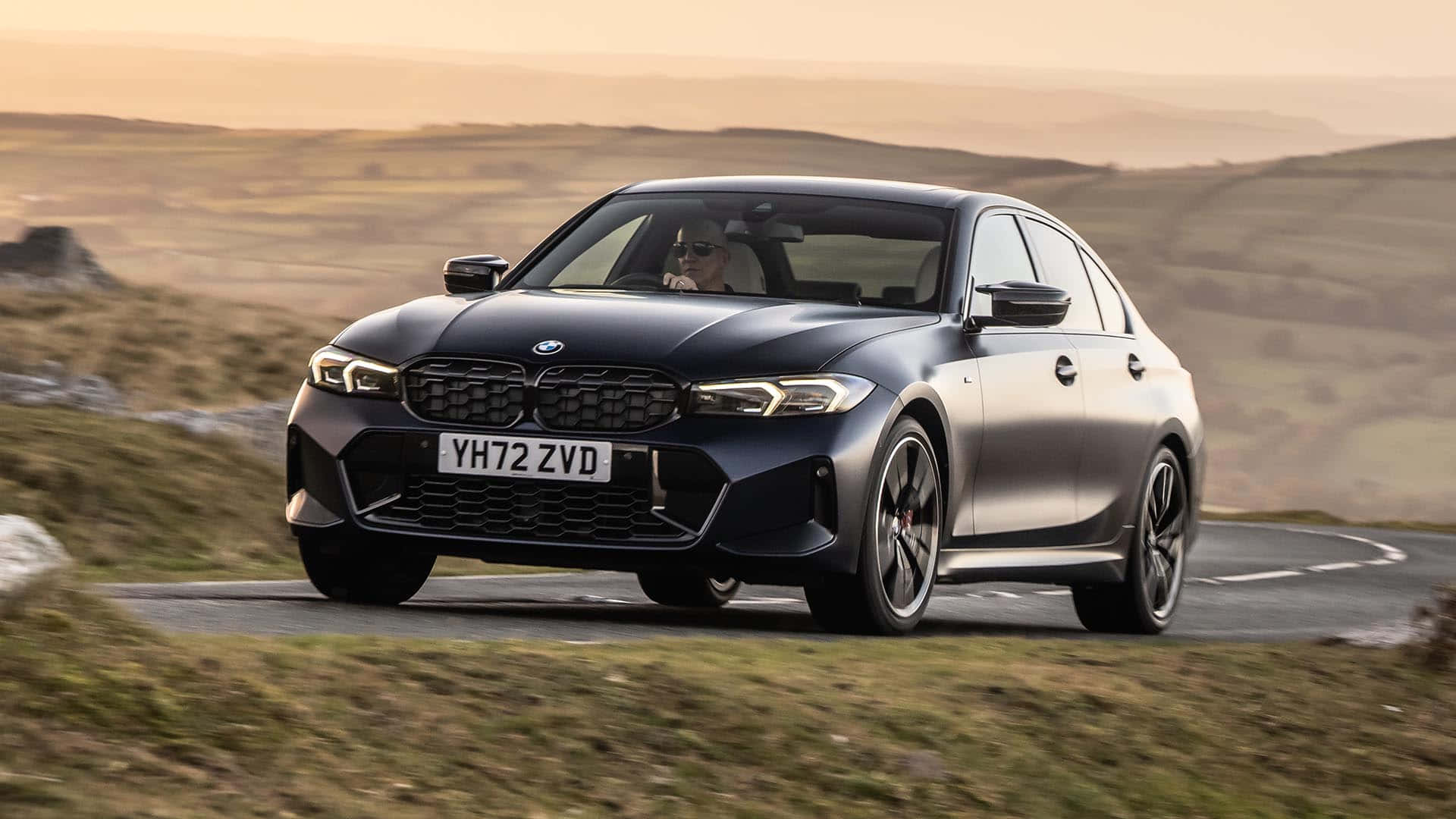 一輛經典BMW賽車在藍色背景中駛過 壁紙 — 一架時尚的啞光黑色BMW M3房車在黃金時段沿著蜿蜒的山路行駛，背景是連綿起伏的山丘。