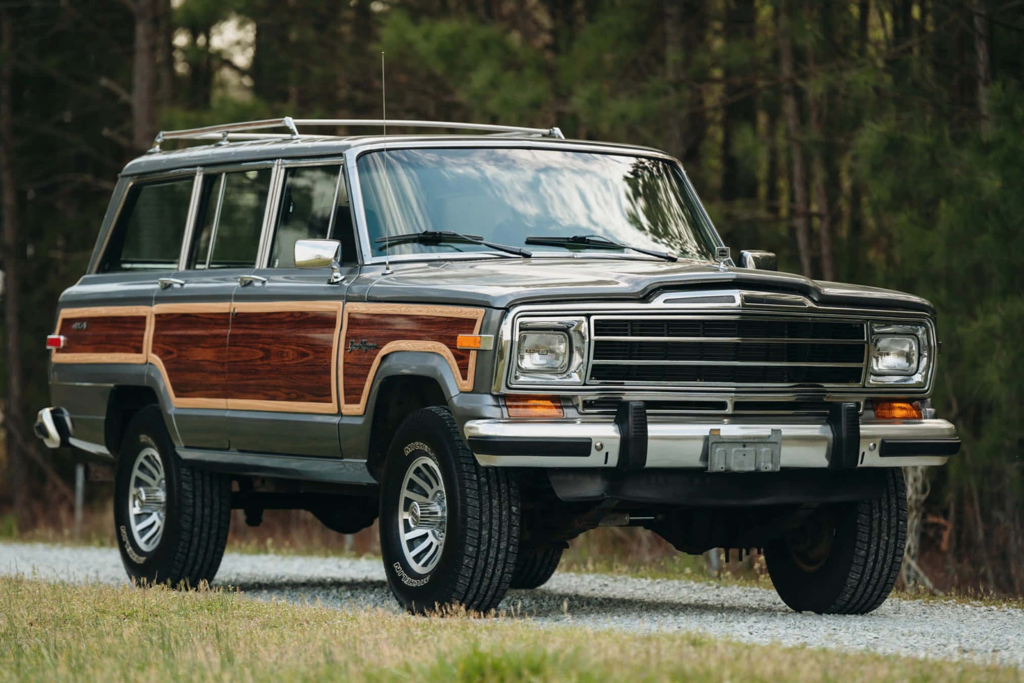 Jeep Wagoneer 桌布 壁紙 — 一輛經典木紋車身Jeep Cherokee旅行車停泊在碎石路上，周圍是高聳的松樹。