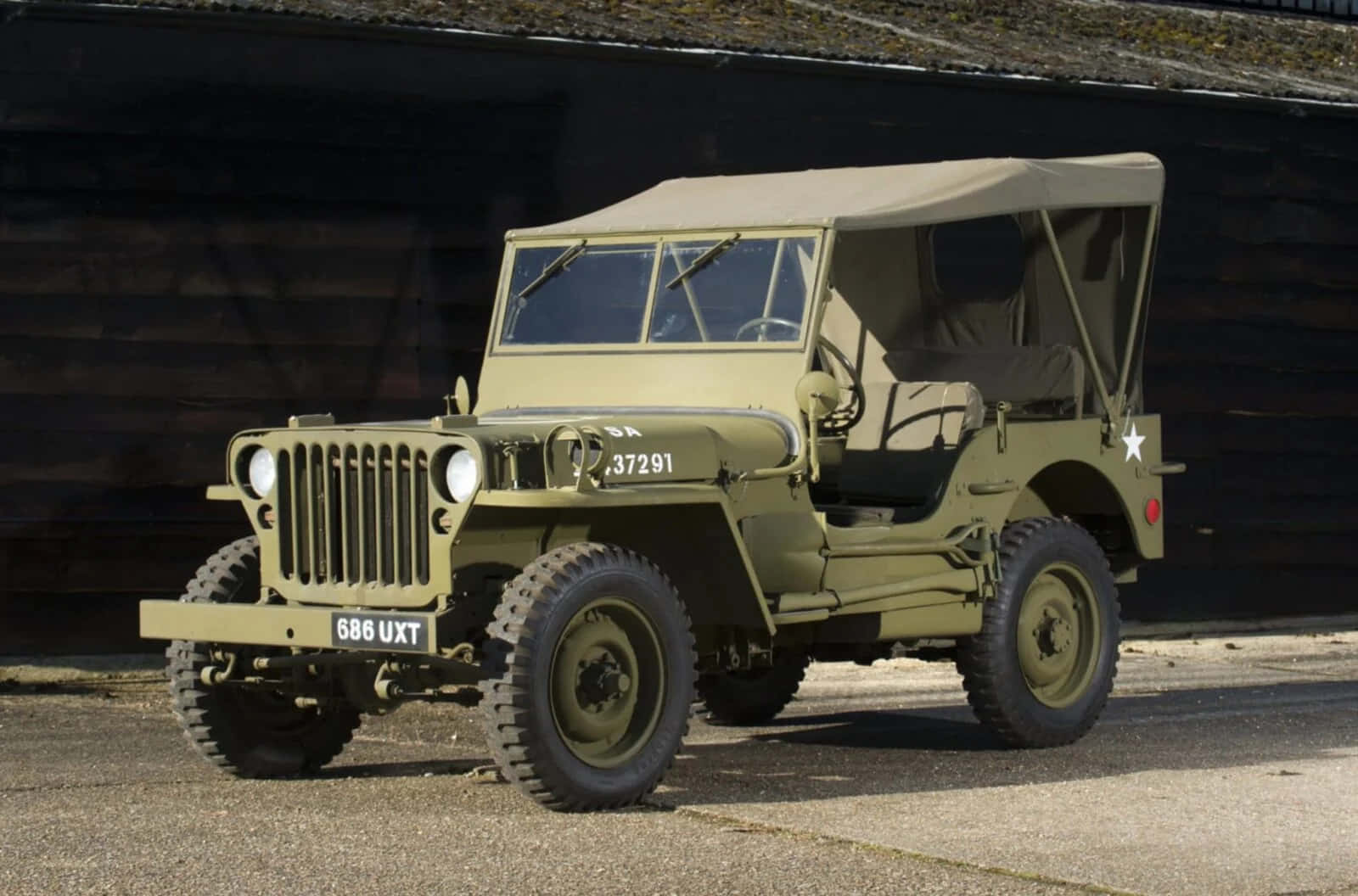 Jeep Willys 桌布 壁紙 — 一輛經典橄欖綠軍用吉普車，帶帆布頂篷，停在瀝青路面上，背景是深色木牆。