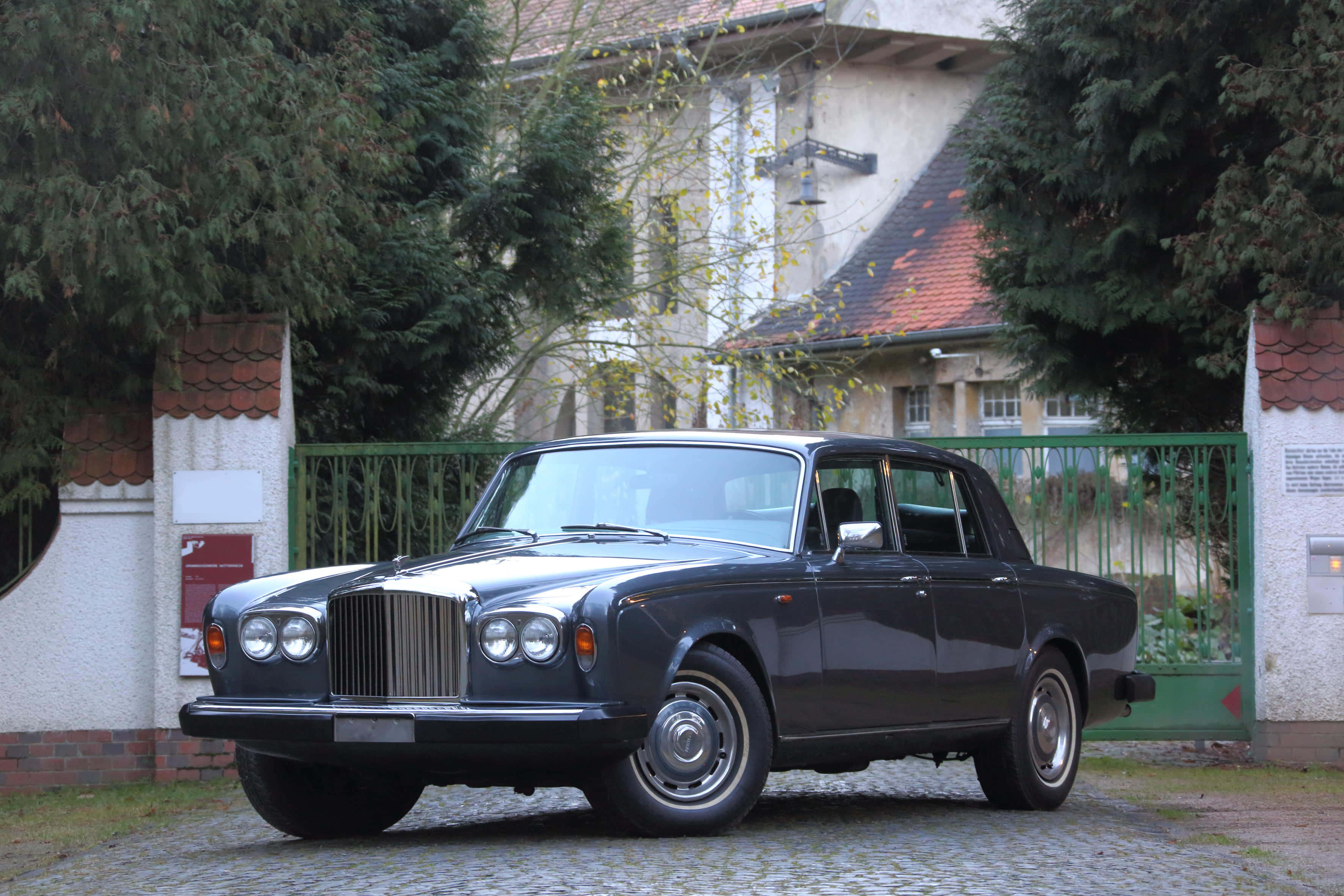 Bentley T2 桌布 壁紙 — 一輛經典深藍色勞斯萊斯優雅地停在鵝卵石車道上，周圍環繞著綠植和復古大門。