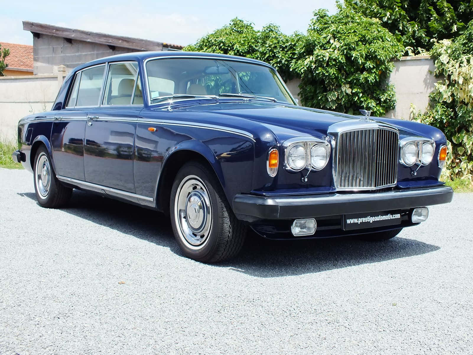 Bentley T2 桌布 壁紙 — 一輛經典深藍色賓利轎車在明亮陽光下閃閃發光，停在碎石路上，旁邊是茂密的綠植。