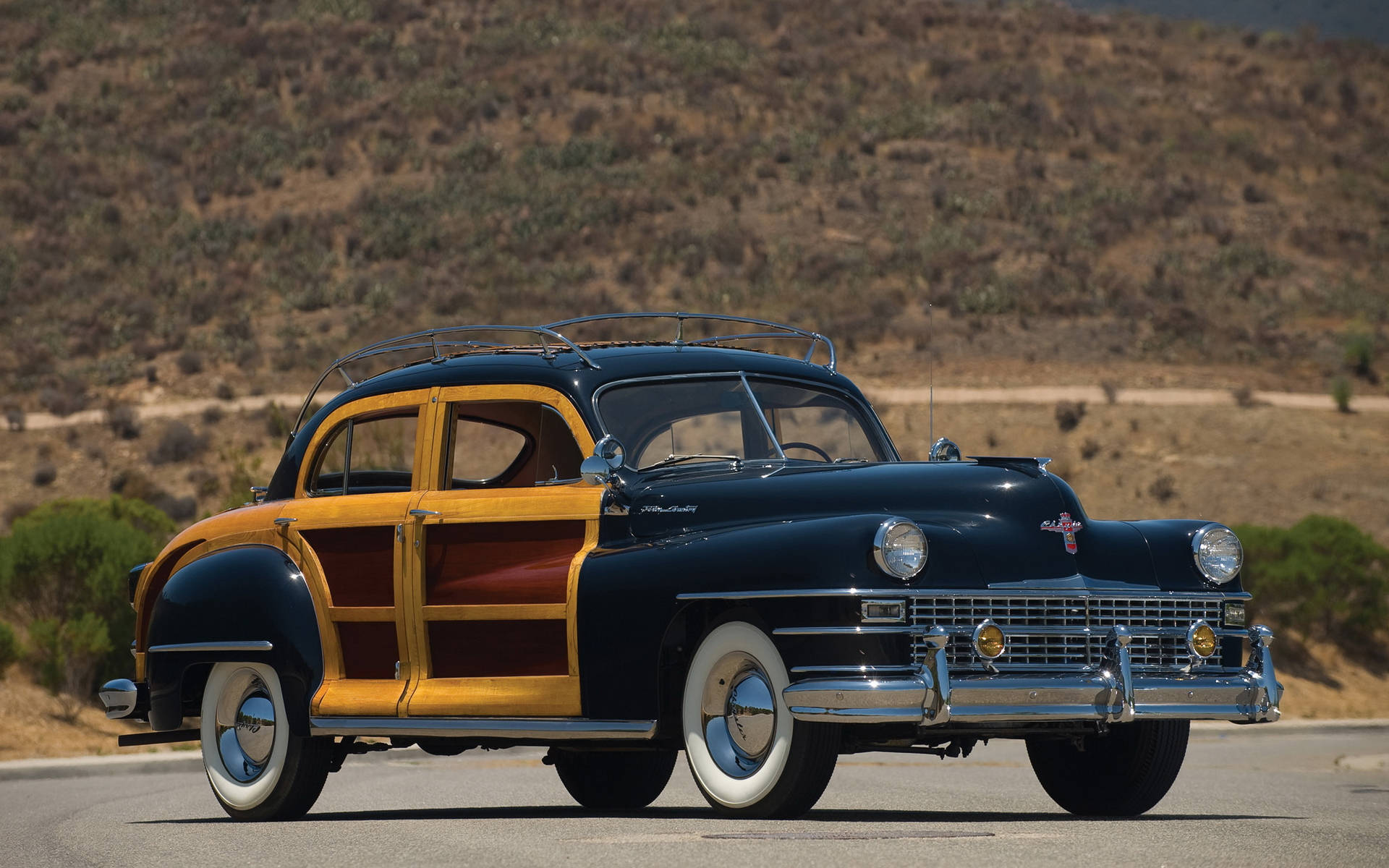 1948 Chrysler Town 壁紙 — 一輛經典的1940年代木鑲板轎車在明亮的陽光下閃耀，行駛在沙漠高速公路上，背景是起伏的山丘。