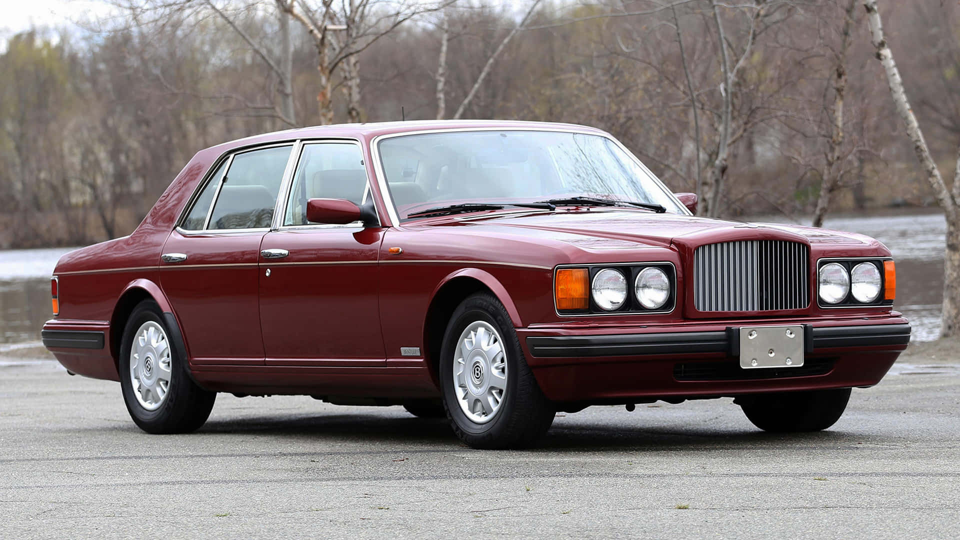 Bentley Brooklands 桌布 壁紙 — 一輛經典的栗色賓利轎車在柔和日光下閃耀，停在柏油路上，旁邊是一條平靜的河流，背景有光禿禿的樹木。