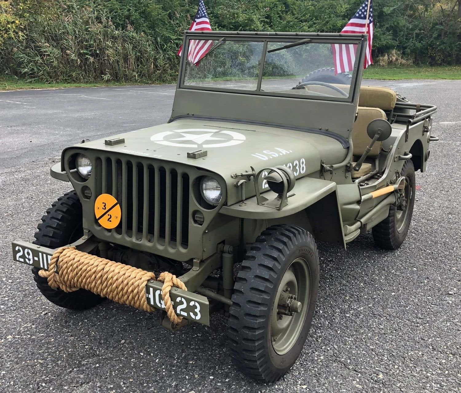 Jeep Willys 桌布 壁紙 — 一輛經典的橄欖綠軍用吉普車，帶有美國國旗和繩索保險槓，停在柏油路上，靠近綠色植物。