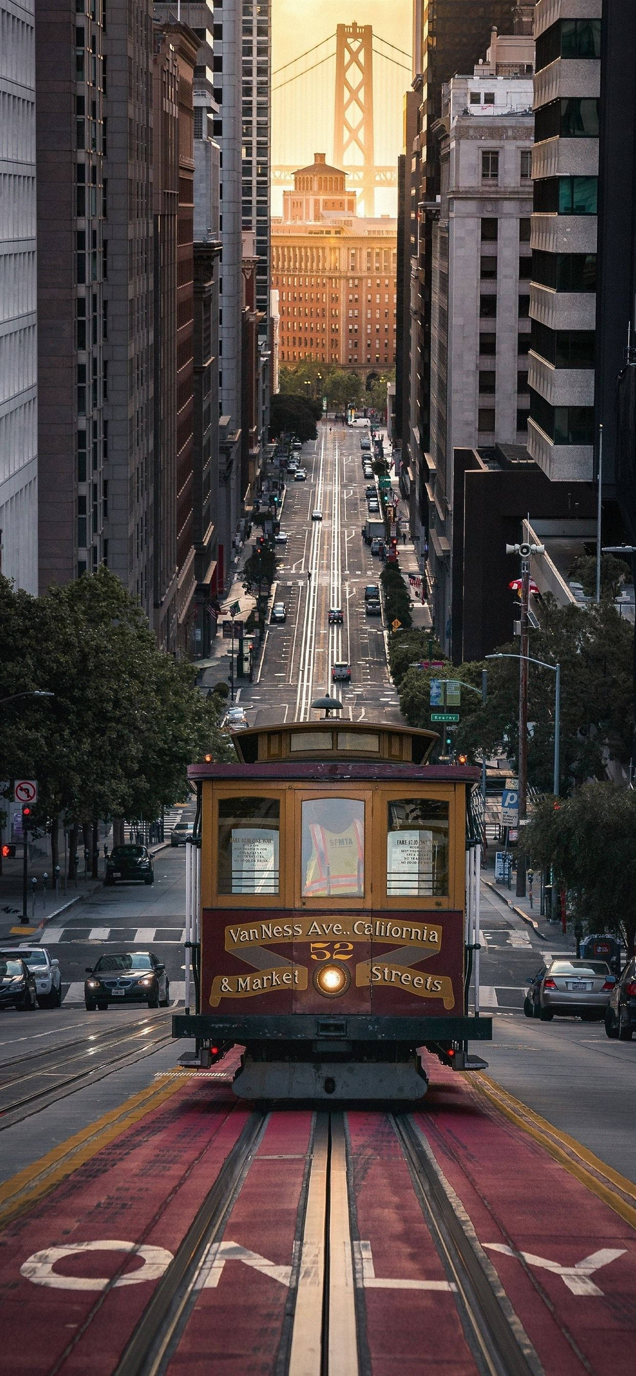San Francisco Phone 桌布 壁紙 — 一輛經典的舊金山纜車沿著陽光明媚的街道滑行，兩旁是高聳的摩天大樓，遠處的金門大橋閃閃發光。