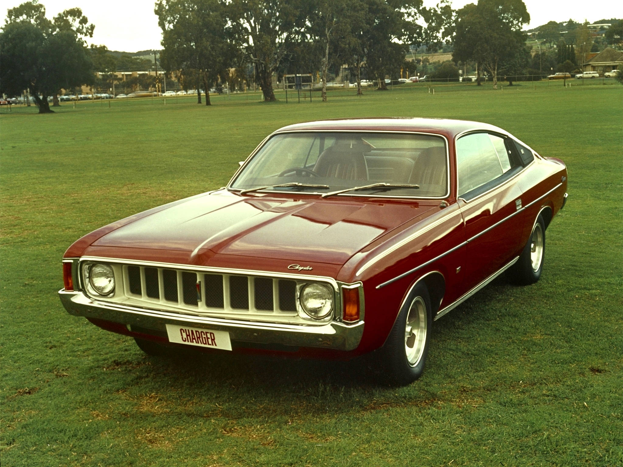 Chrysler Valiant Charger 壁紙 — 一輛經典紅色Chrysler Charger雙門跑車停放在茂密的綠色草坪上，背景有樹木和遠處建築物，在柔和日光下。