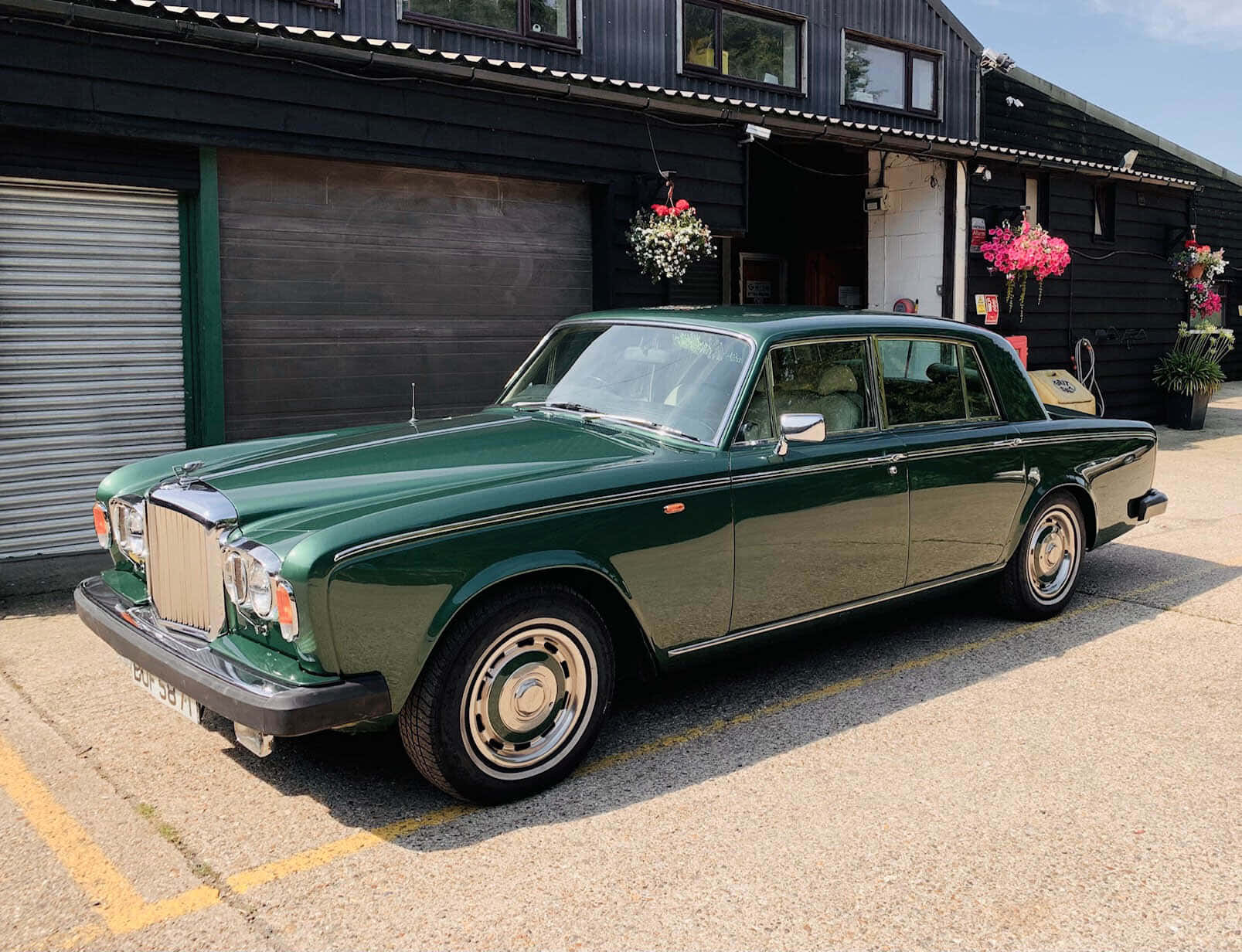 Bentley T2 桌布 壁紙 — 一輛經典綠色勞斯萊斯銀影在明亮陽光下閃閃發光，停放在一個裝飾著懸掛花籃的深色木車庫前。