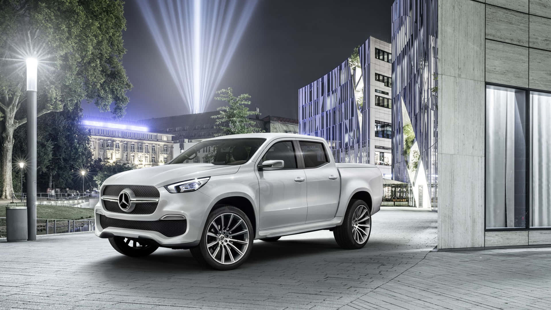 銀色 Mercedes-Benz 皮卡車在夜間停放在現代城市廣場，配有聚光燈和玻璃建築。