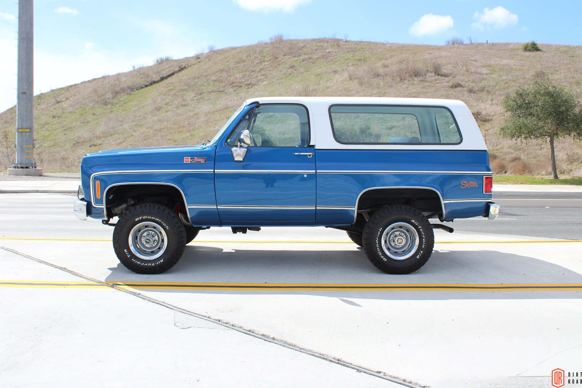 Gmc Jimmy 桌布 壁紙 — 一輛經典藍白色升高底盤的雪佛蘭 K5 Blazer，配備鍍鉻輪圈，停泊在鋪砌道路上，旁邊是乾燥的草坡，上方是明亮的藍天。