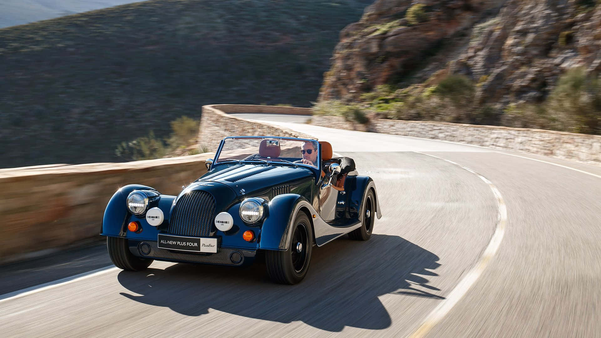 Morgan 桌布 壁紙 — 一輛經典藍色摩根Plus Four開篷跑車在明媚陽光下沿著蜿蜒的山路飛馳。