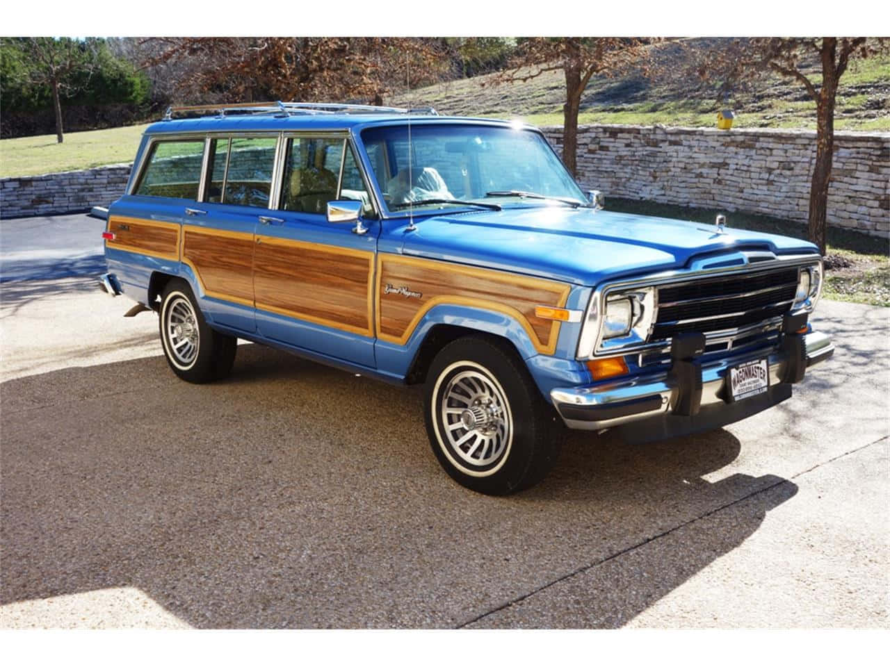 Jeep Wagoneer 桌布 壁紙 — 一輛經典藍色木紋車身Jeep Wagoneer停泊在陽光普照的混凝土車道上，旁邊有石牆和樹木。