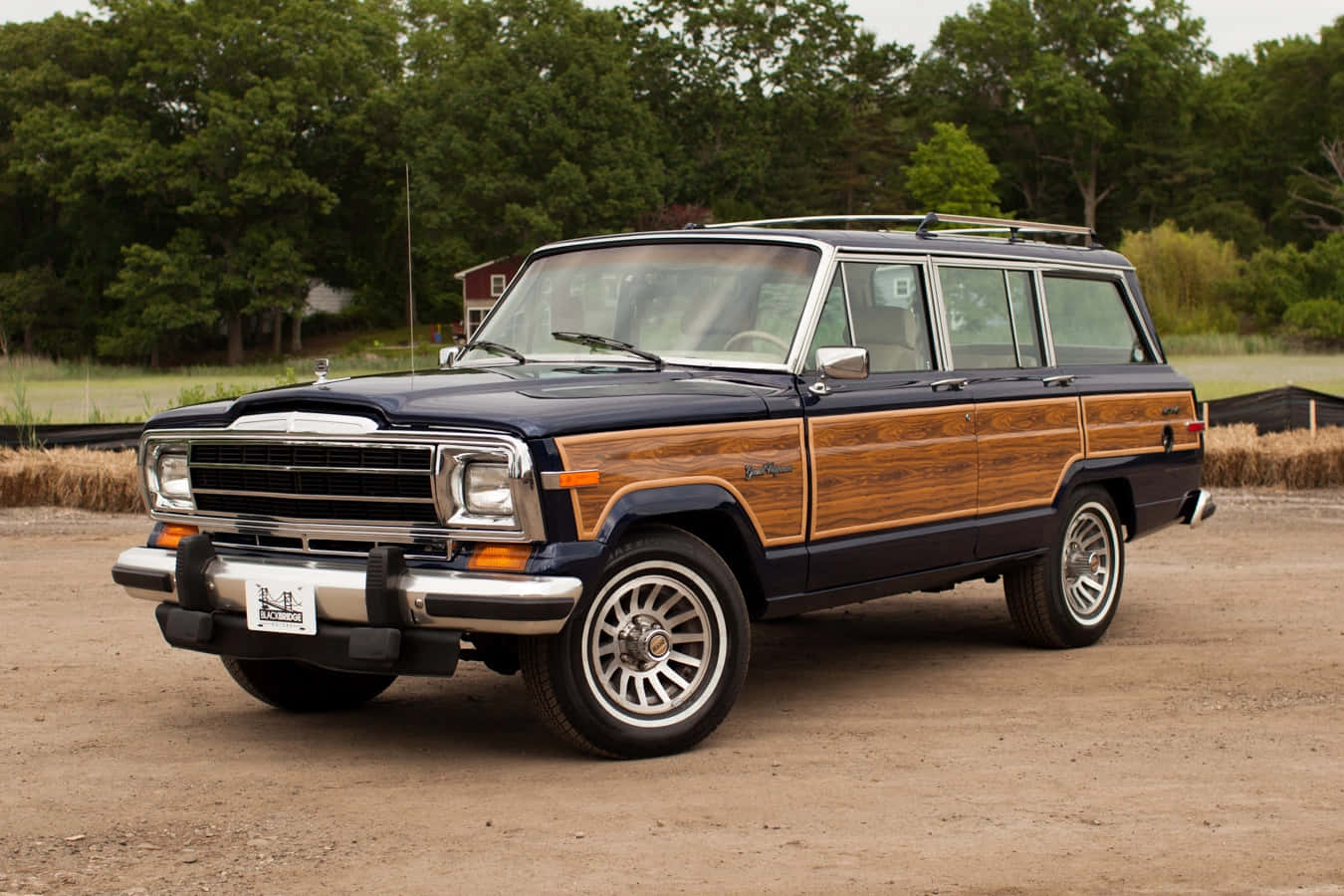 Jeep Wagoneer 桌布 壁紙 — 一輛經典藍色木紋車身旅行車停泊在泥地上，背景有樹木和一個紅色穀倉。