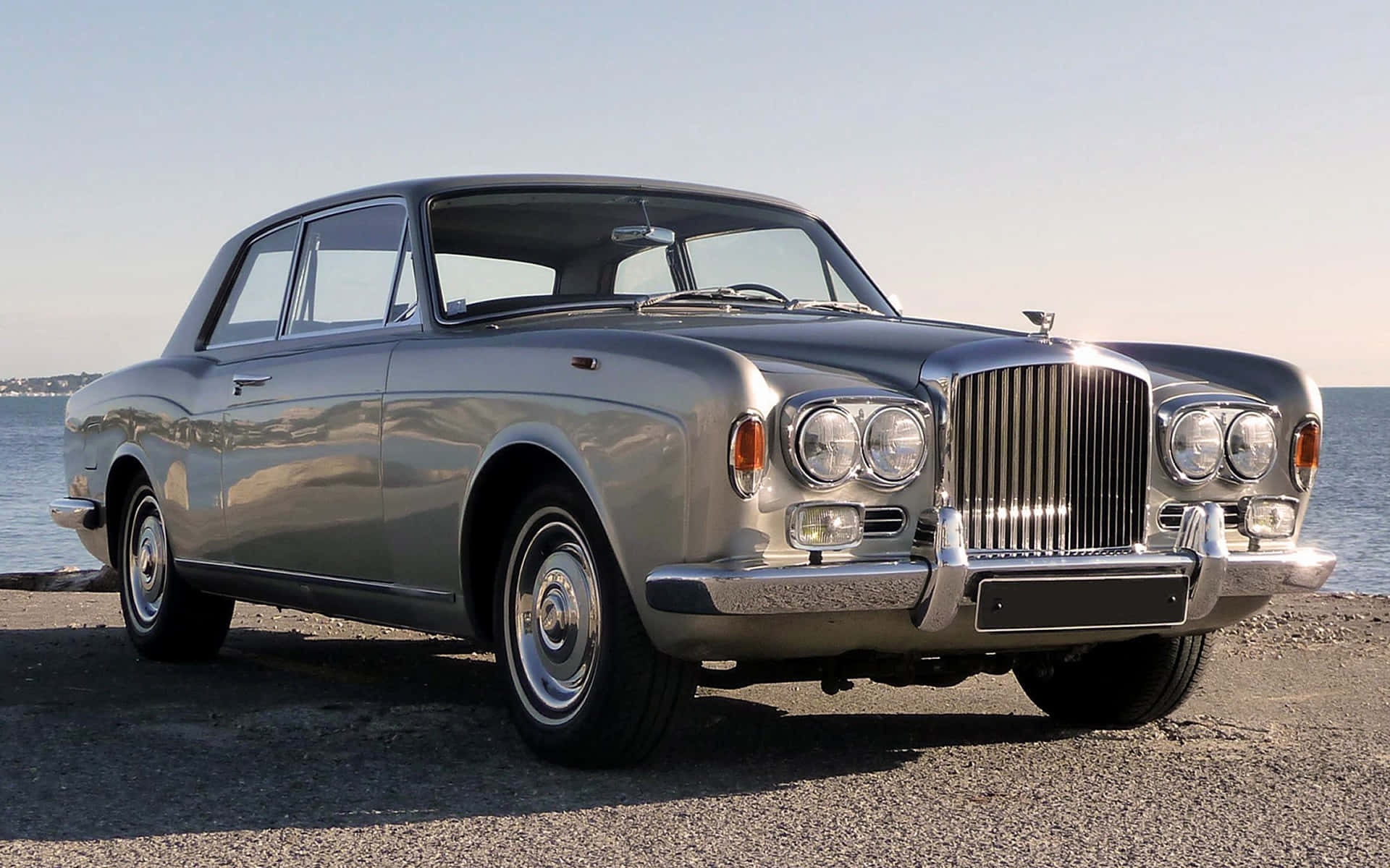 Bentley T2 桌布 壁紙 — 一輛經典銀色賓利轎車在明亮陽光下閃閃發光，停在平靜藍色水邊的礫石岸上。