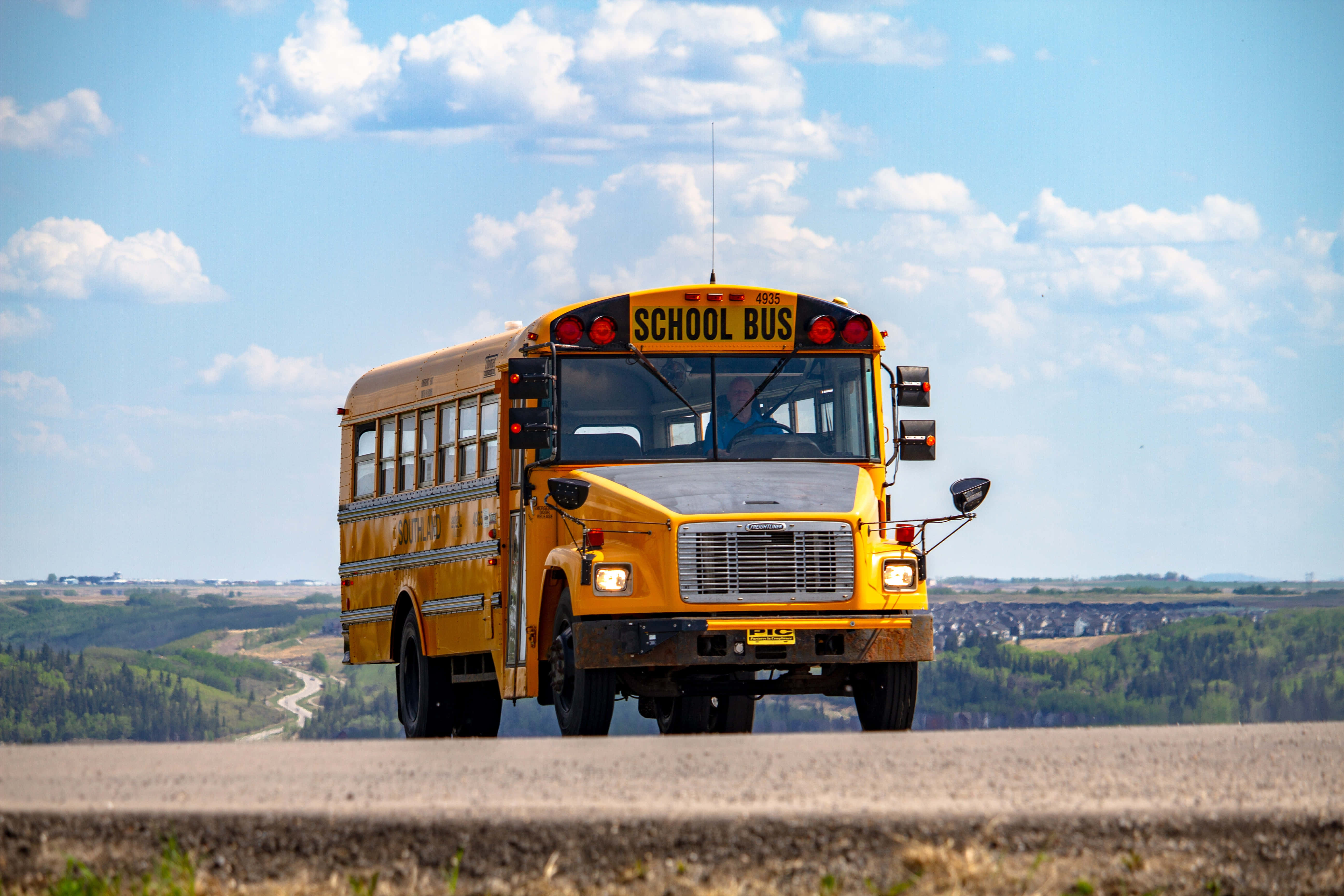 Yellow School Bus 桌布 壁紙 — 一輛經典黃色校車在鄉村道路上向鏡頭駛來，天空湛藍，白雲朵朵。