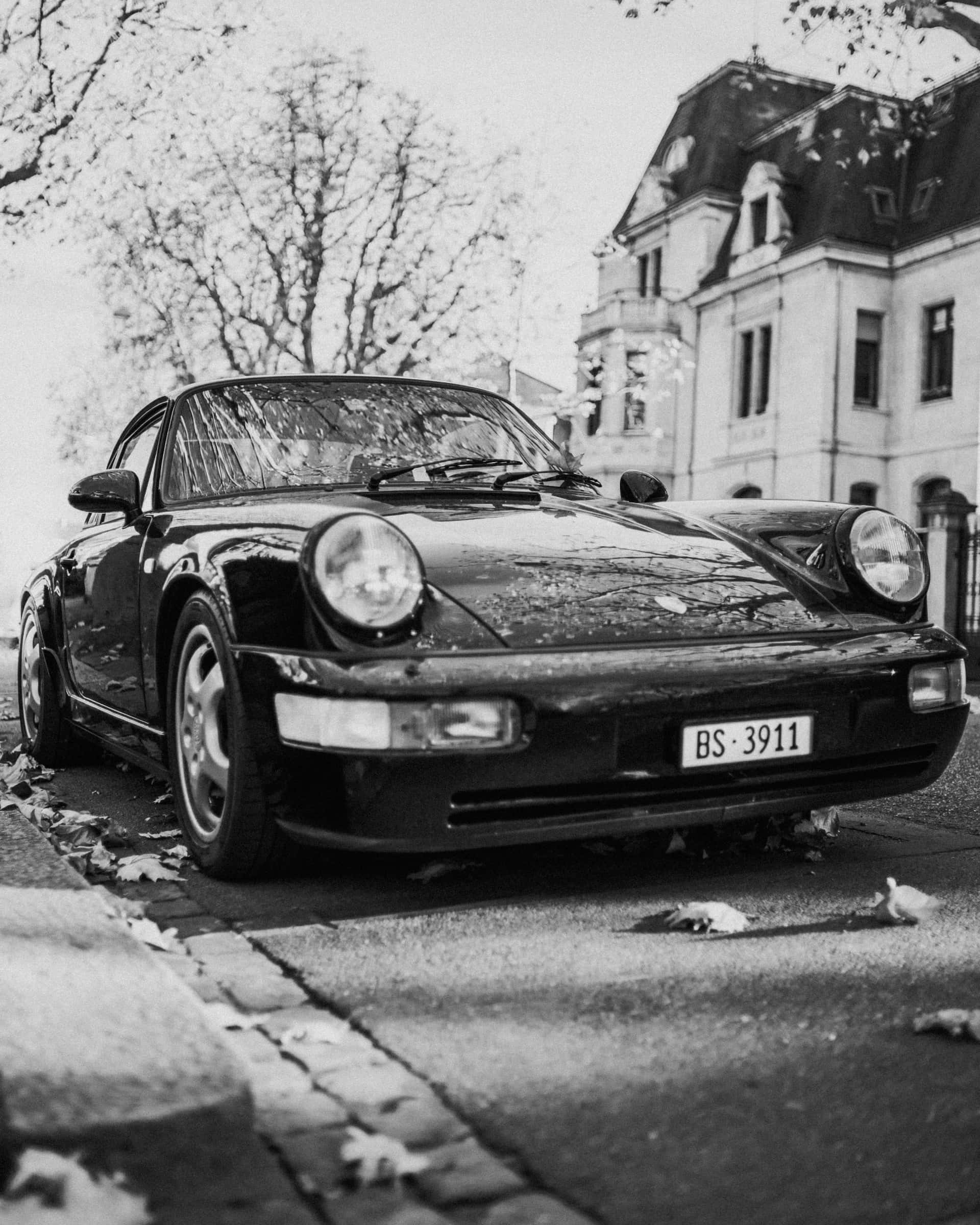 Black And White Car 桌布 壁紙 — 一輛經典黑色保時捷911停在鵝卵石街道上，街道上散落著落葉，背景是一座宏偉的建築。