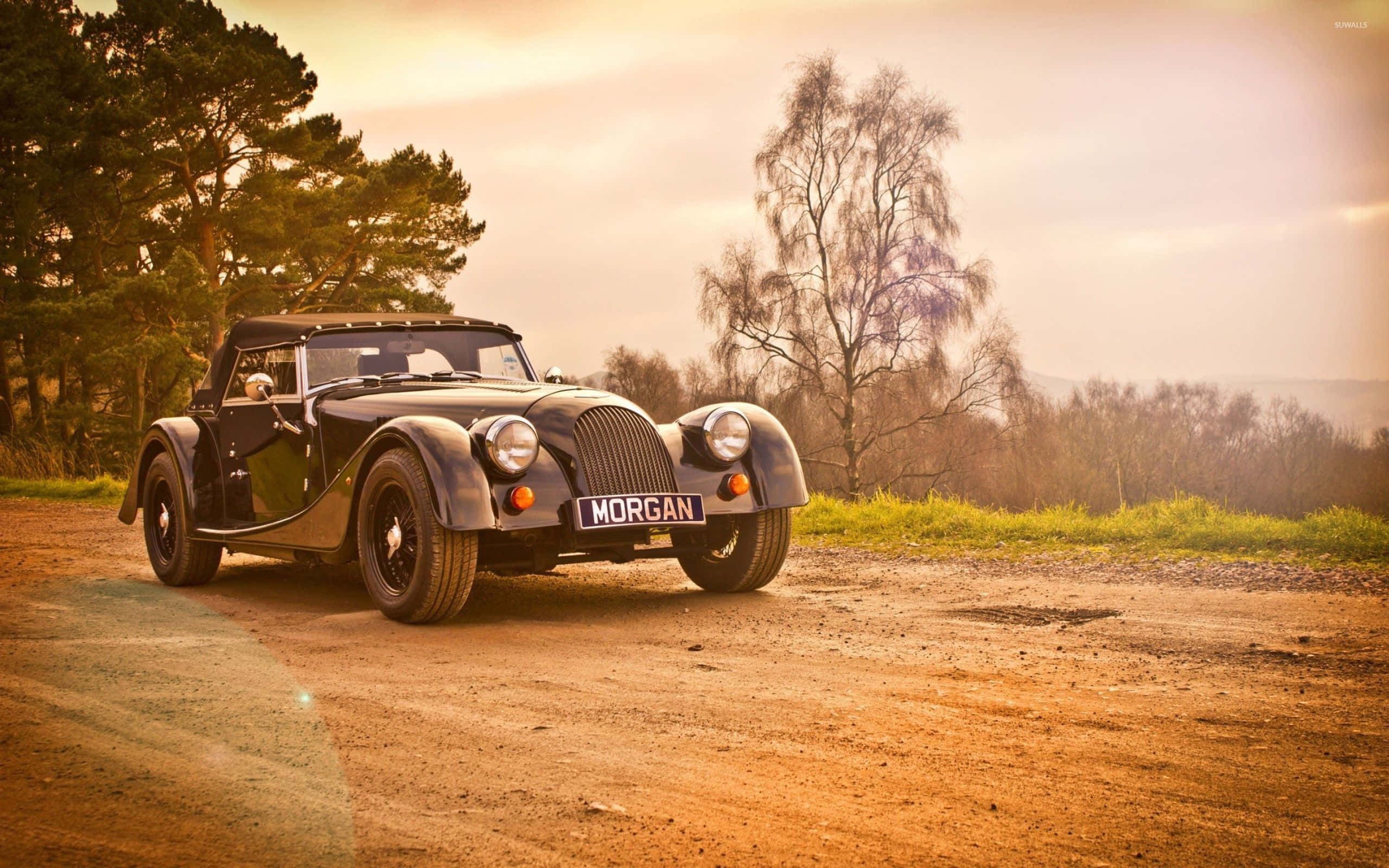 Morgan 桌布 壁紙 — 一輛經典黑色摩根跑車停泊在泥路上，背景是黃金時段的樹木和山丘。