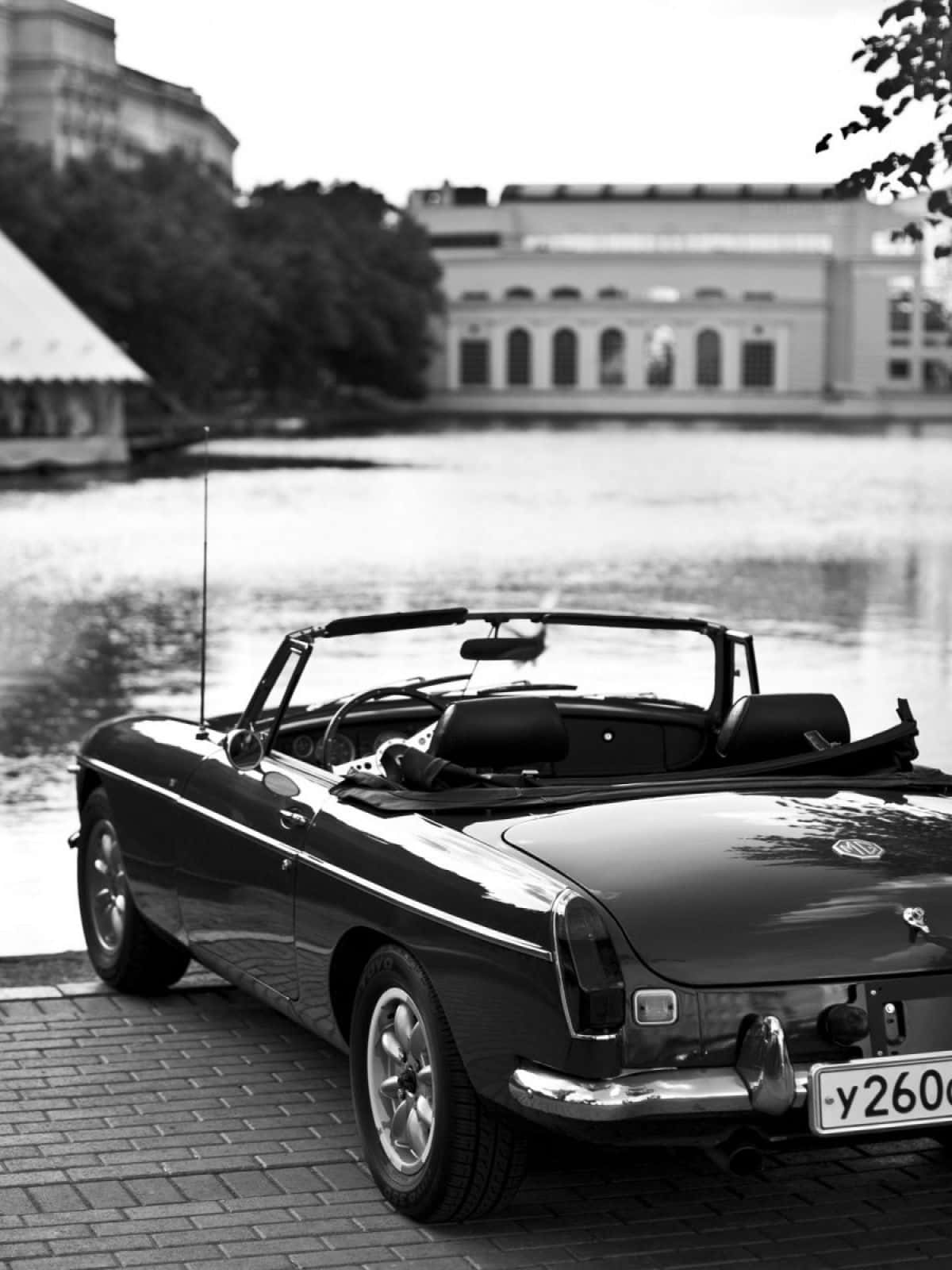 Black And White Car 桌布 壁紙 — 一輛經典黑色開篷車停泊在磚砌碼頭旁，旁邊是平靜的水面，背景是歷史建築。
