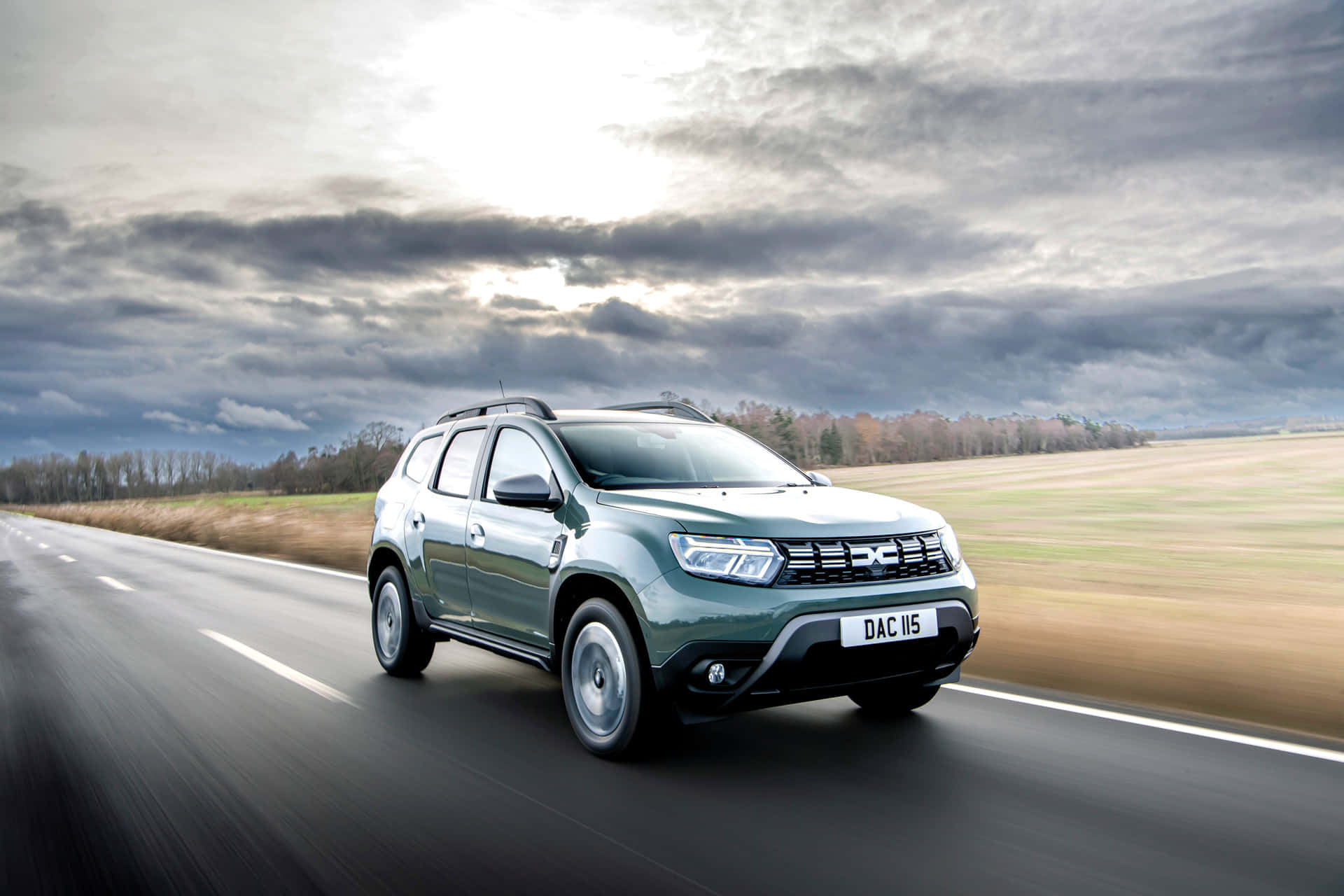 Dacia Duster 桌布 壁紙 — 一輛綠色Dacia Duster SUV在戲劇性的多雲天空下沿鄉村道路飛馳，背景是模糊的田野。
