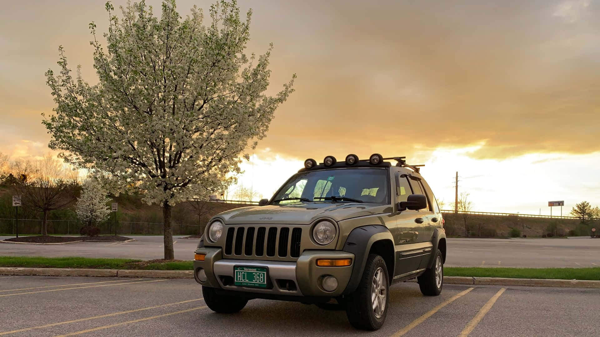 Jeep Liberty 桌布 壁紙 — 一輛綠色Jeep Liberty停泊在停車場，旁邊有一棵盛開的樹，背景是金色的日落天空。