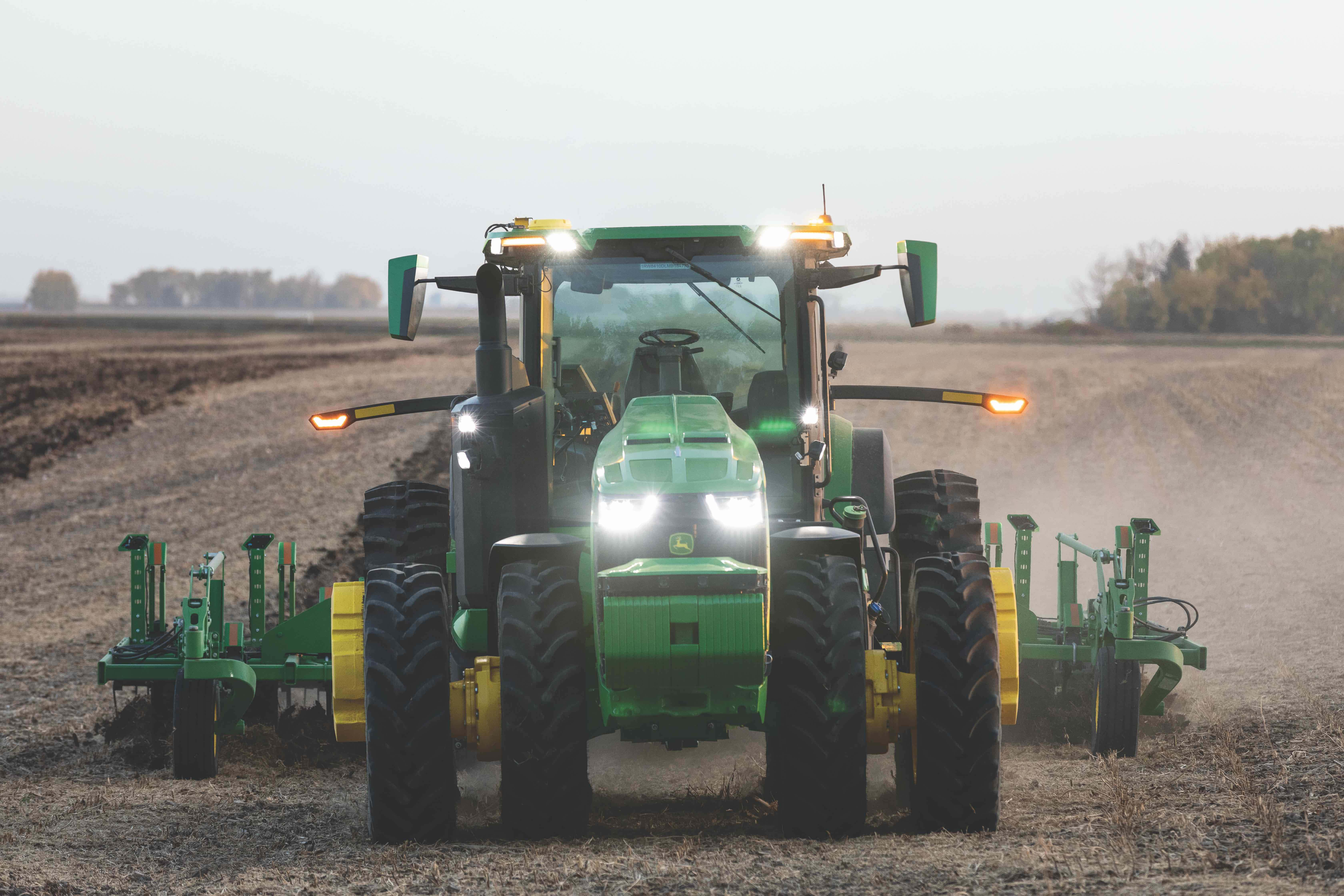 John Deere 桌布 壁紙 — 一輛綠色約翰迪爾拖拉機，配有明亮的頭燈，在黎明時分犁過廣闊的塵土飛揚的田野。