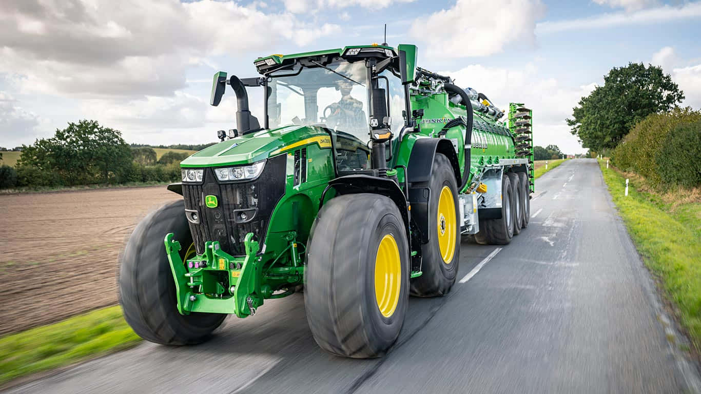 John Deere 桌布 壁紙 — 一輛綠色約翰迪爾拖拉機，黃色車輪，拖著拖車行駛在田野和樹木環繞的鄉村道路上，天空多雲。