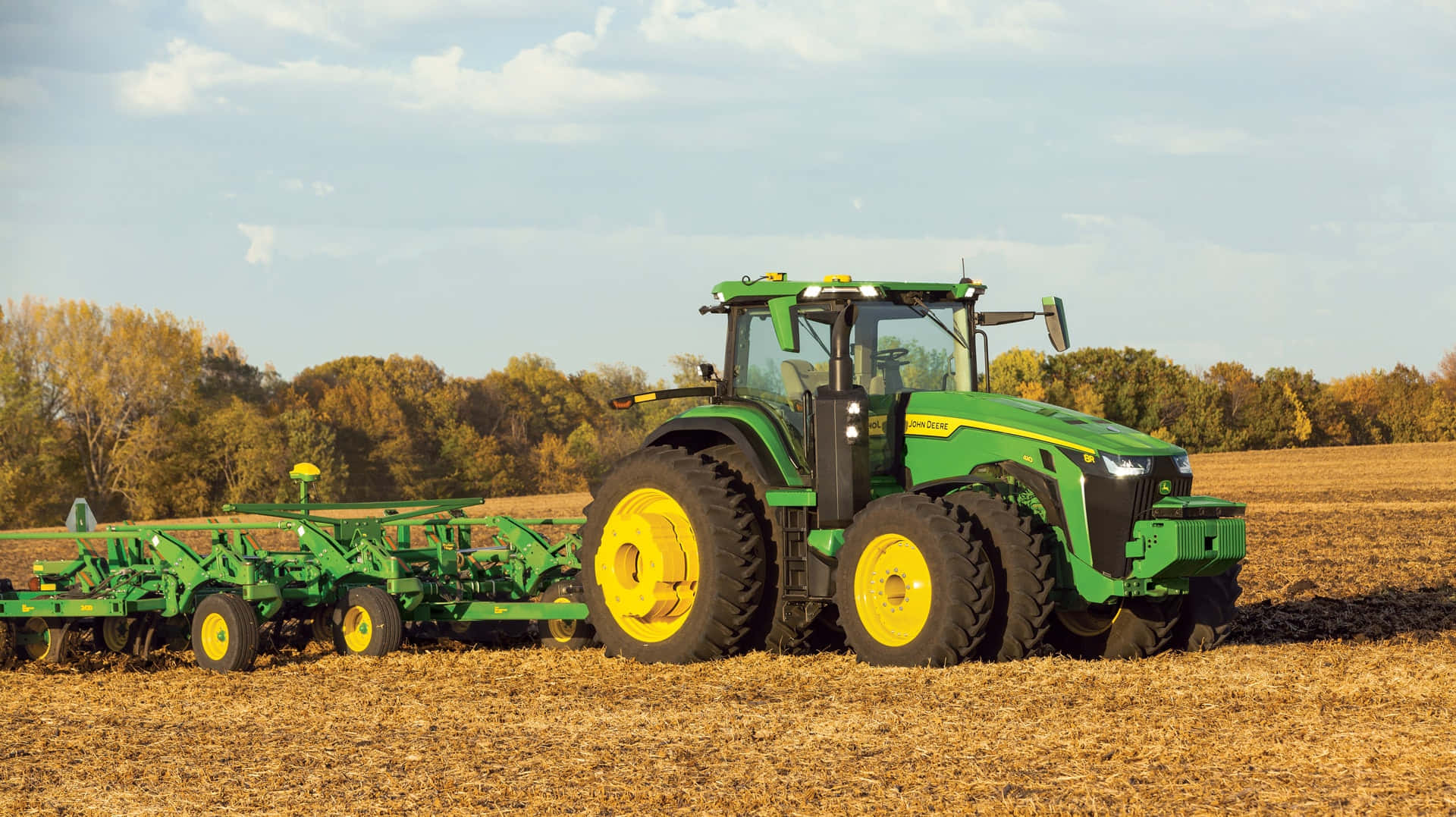 John Deere 桌布 壁紙 — 一輛綠色約翰迪爾拖拉機，黃色車輪，在部分多雲的天空下，拖著犁穿過金色的收割過的田野。